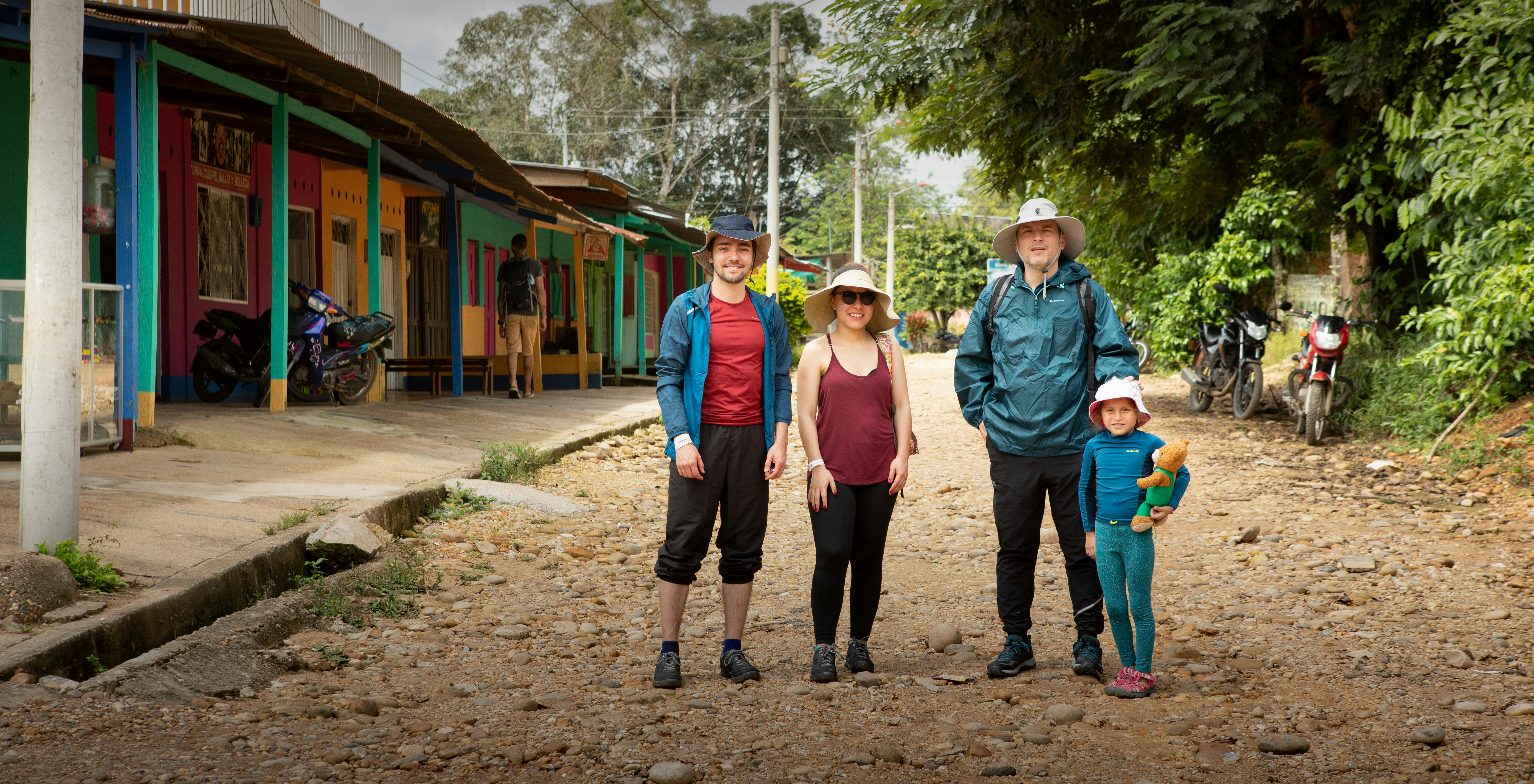 Three adults and a little girl in blue rash guards who is holding a teddy bear smile for the camera in front of a colorful row of shops and other businesses on a rocky dirt road in Colombia