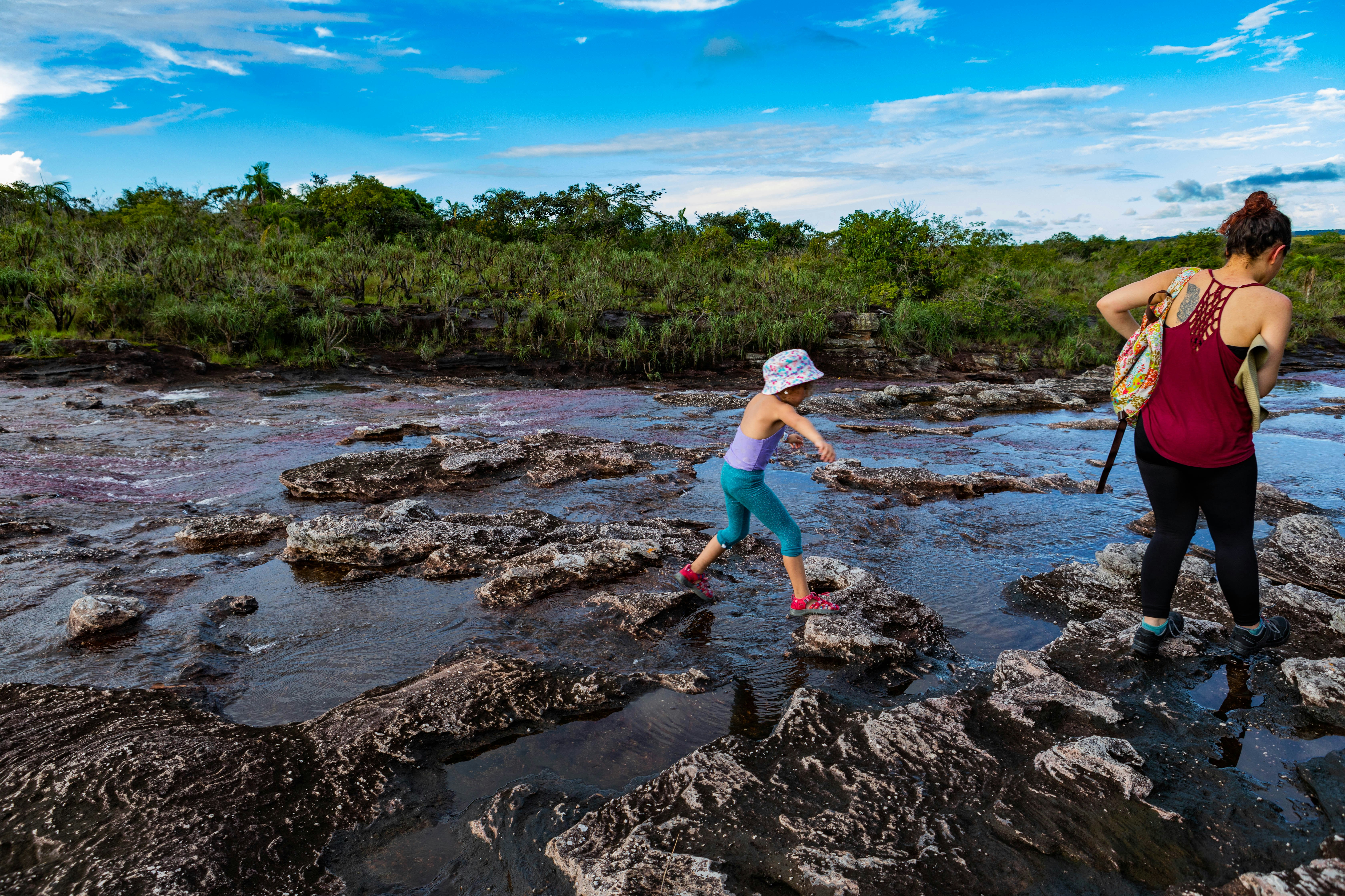 A little girl in blue leggings and a purple swim top hops between rocks on the Cano Cristales river next to a woman in a red tank top and black leggings