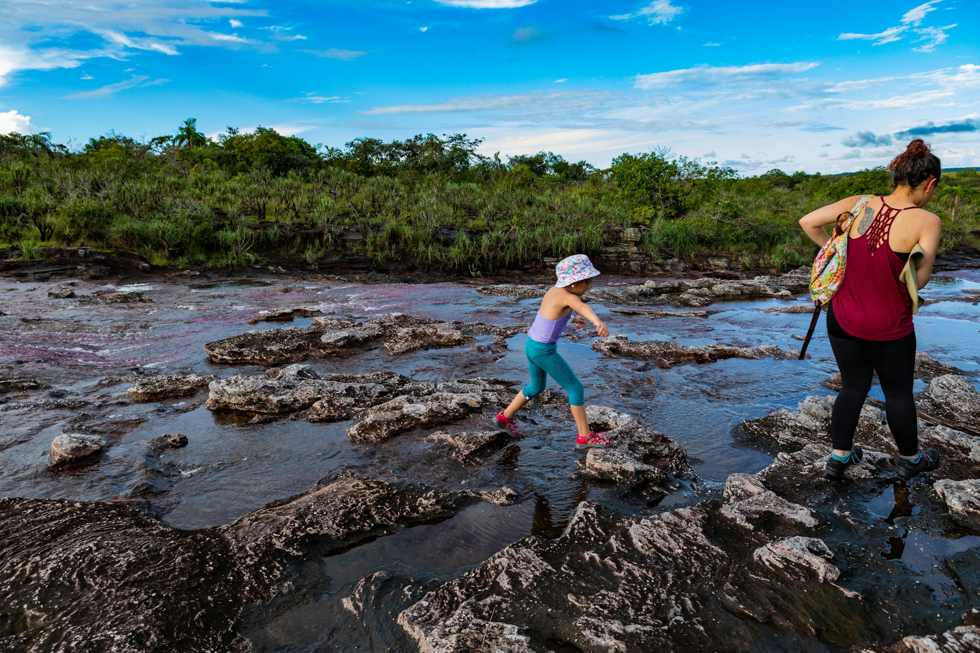 A little girl in blue leggings and a purple swim top hops between rocks on the Cano Cristales river next to a woman in a red tank top and black leggings