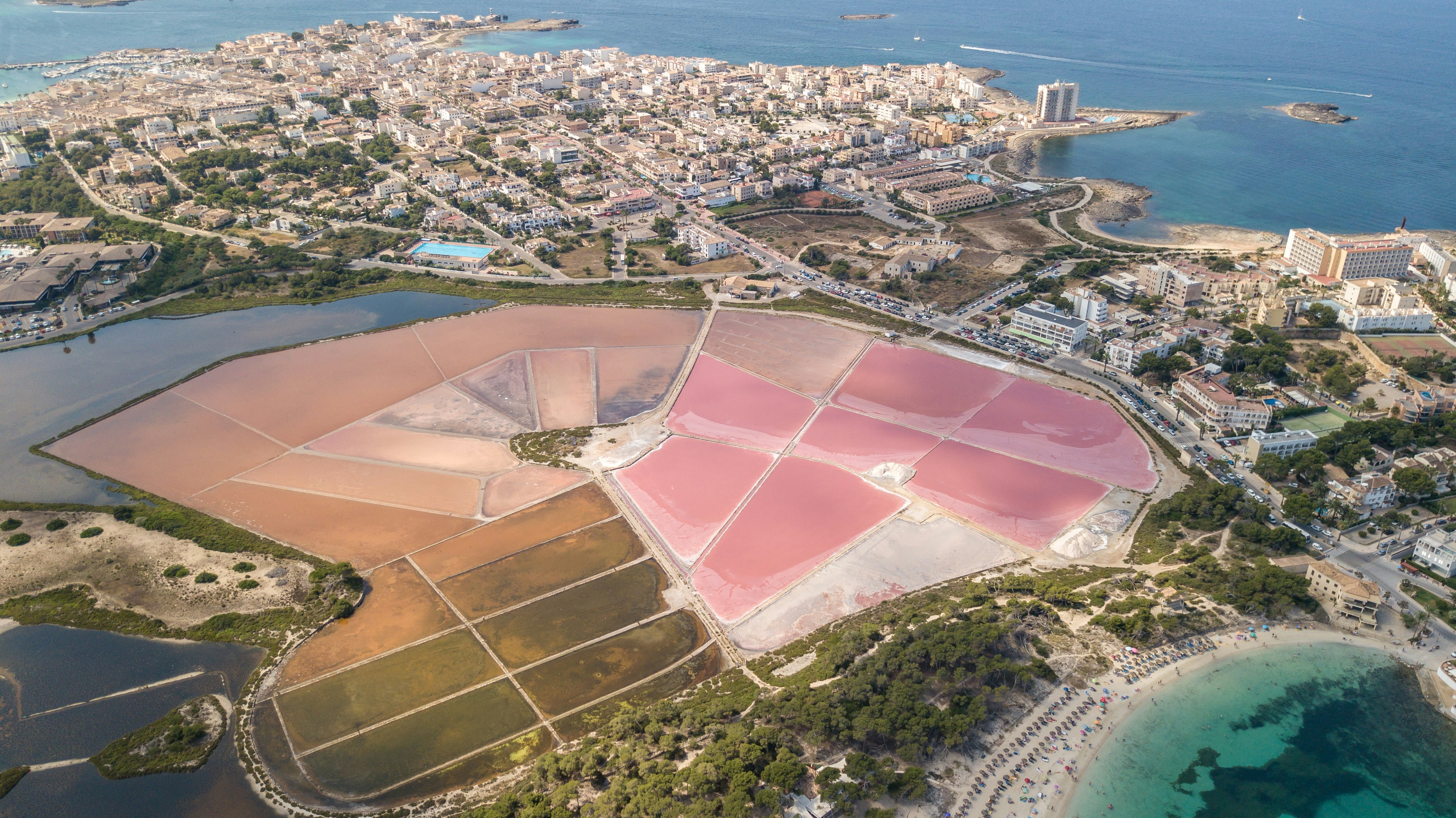 Pink salt flats sit near a turquoise lagoon and blue sea waters rimmed by a white cityscape at Colonia de Sant Jordi in Spain