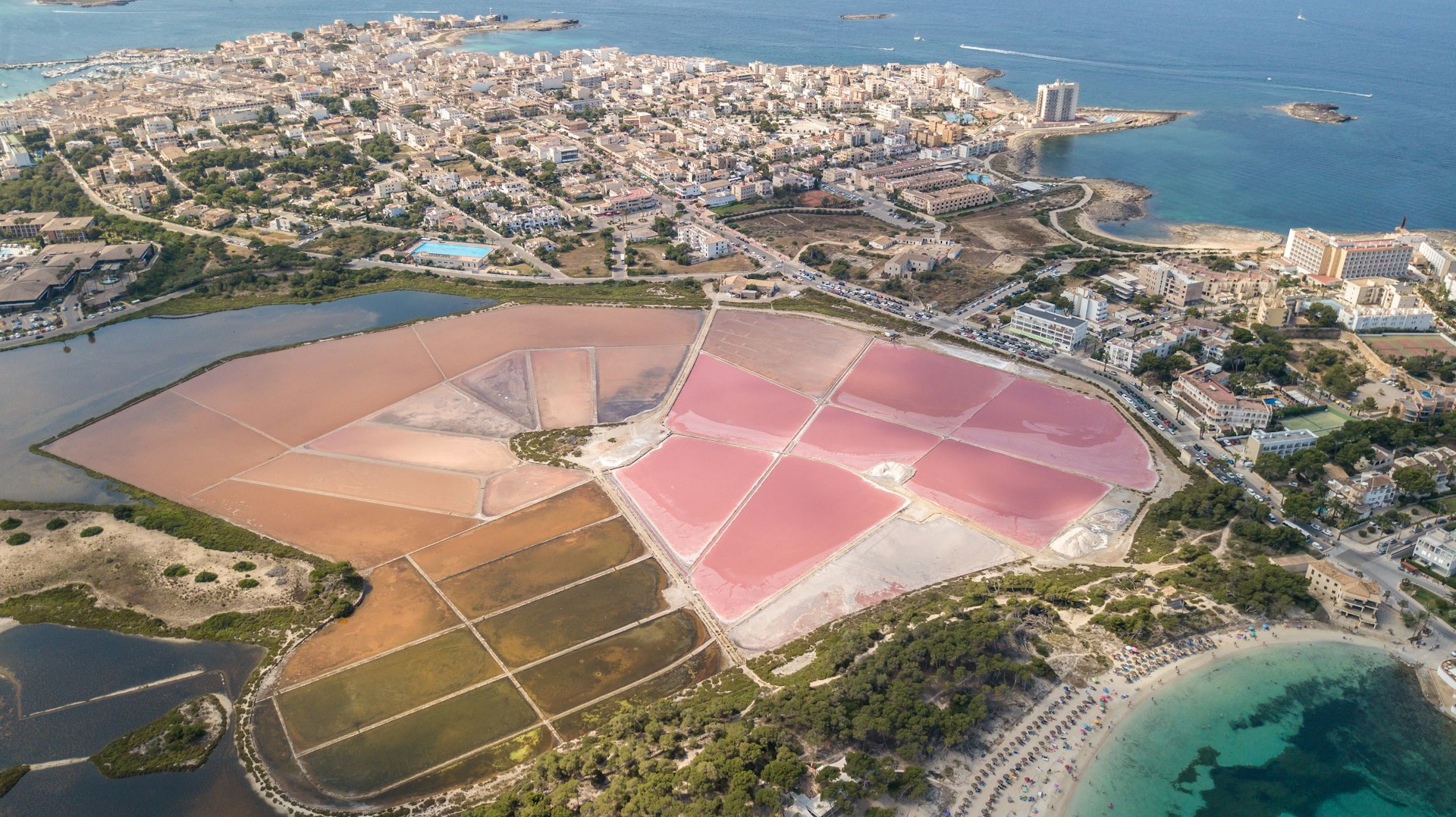 Pink salt flats sit near a turquoise lagoon and blue sea waters rimmed by a white cityscape at Colonia de Sant Jordi in Spain