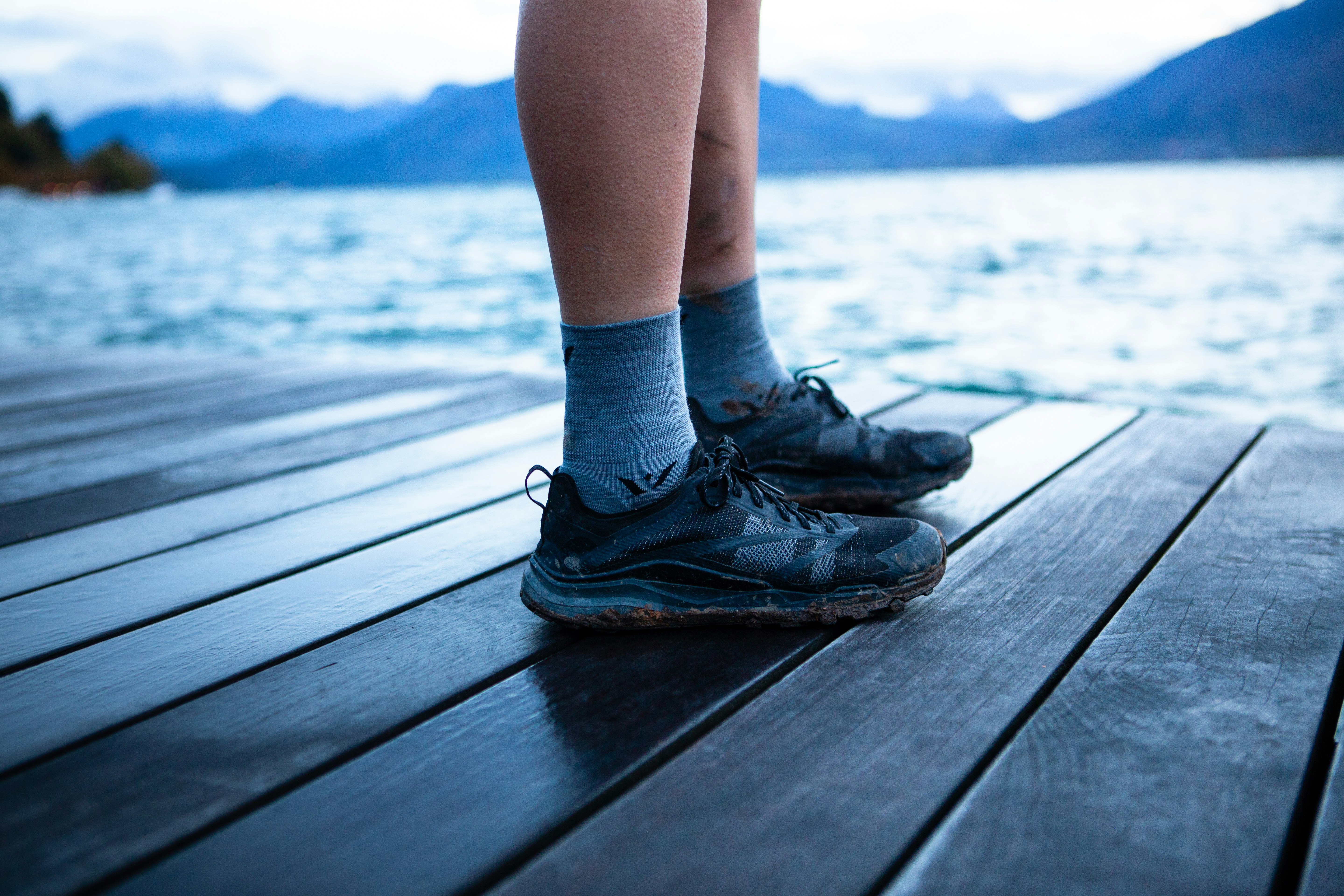 Comfy socks and muddy trail shoes stand on a dock near a lake