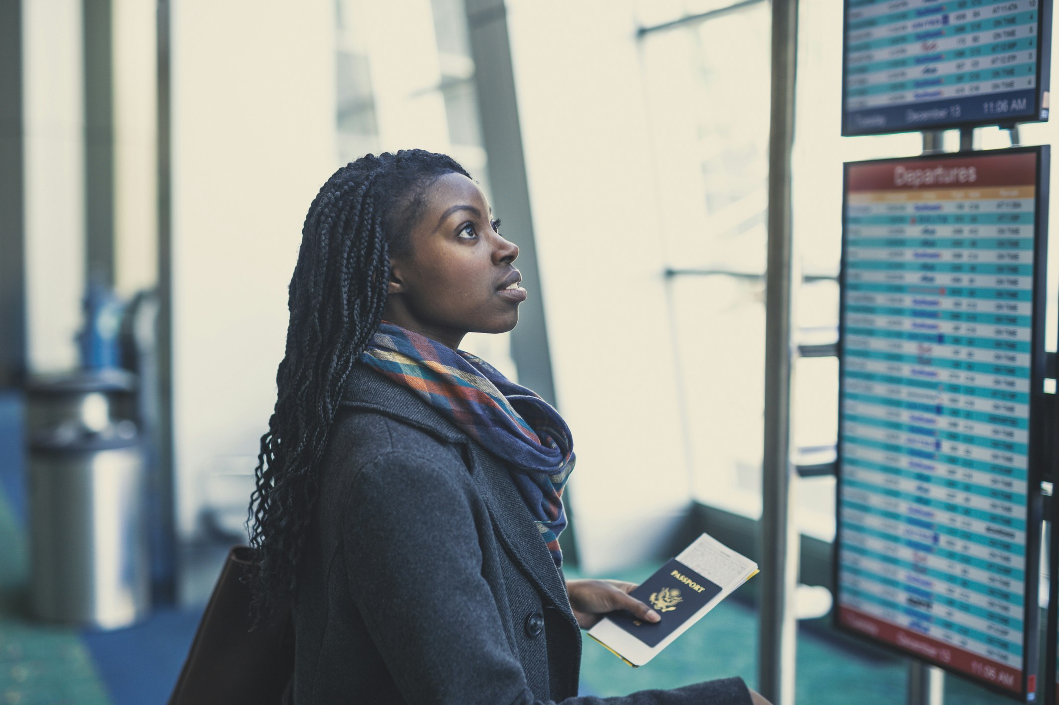 An African-American woman with long braids and a blue blazer looks up hopefully at a departures board in an airport. She is holding a passport and her tickets in her left hand