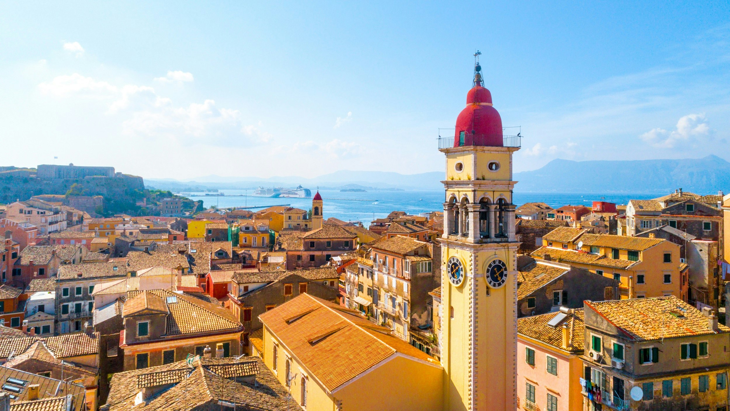 Yellow brick buildings and rooftops, with a large clock tower with a red roof. The sea is in the distance, with hills beyond.