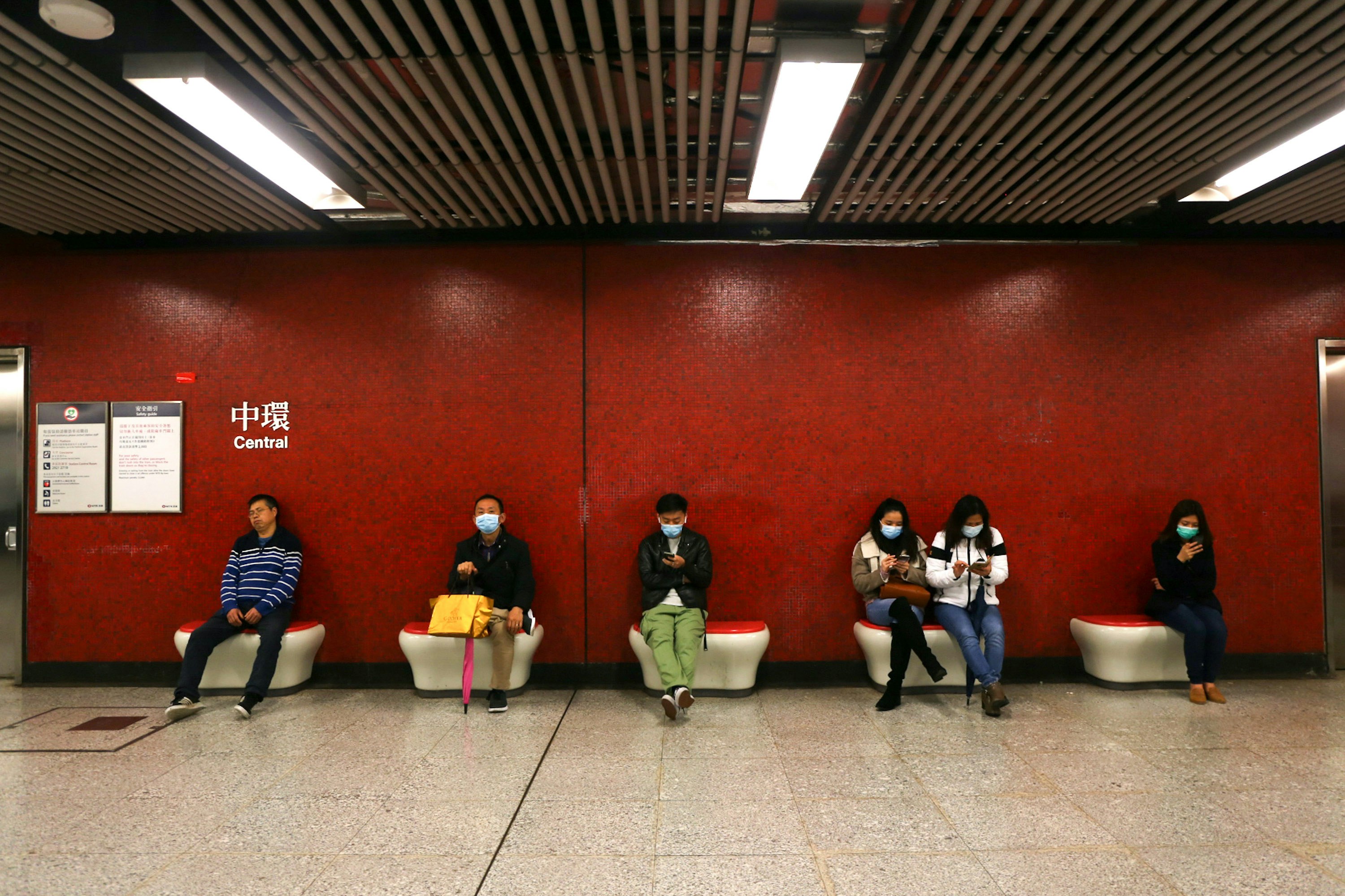 People sit wearing face masks in front a red wall in a subway station. 