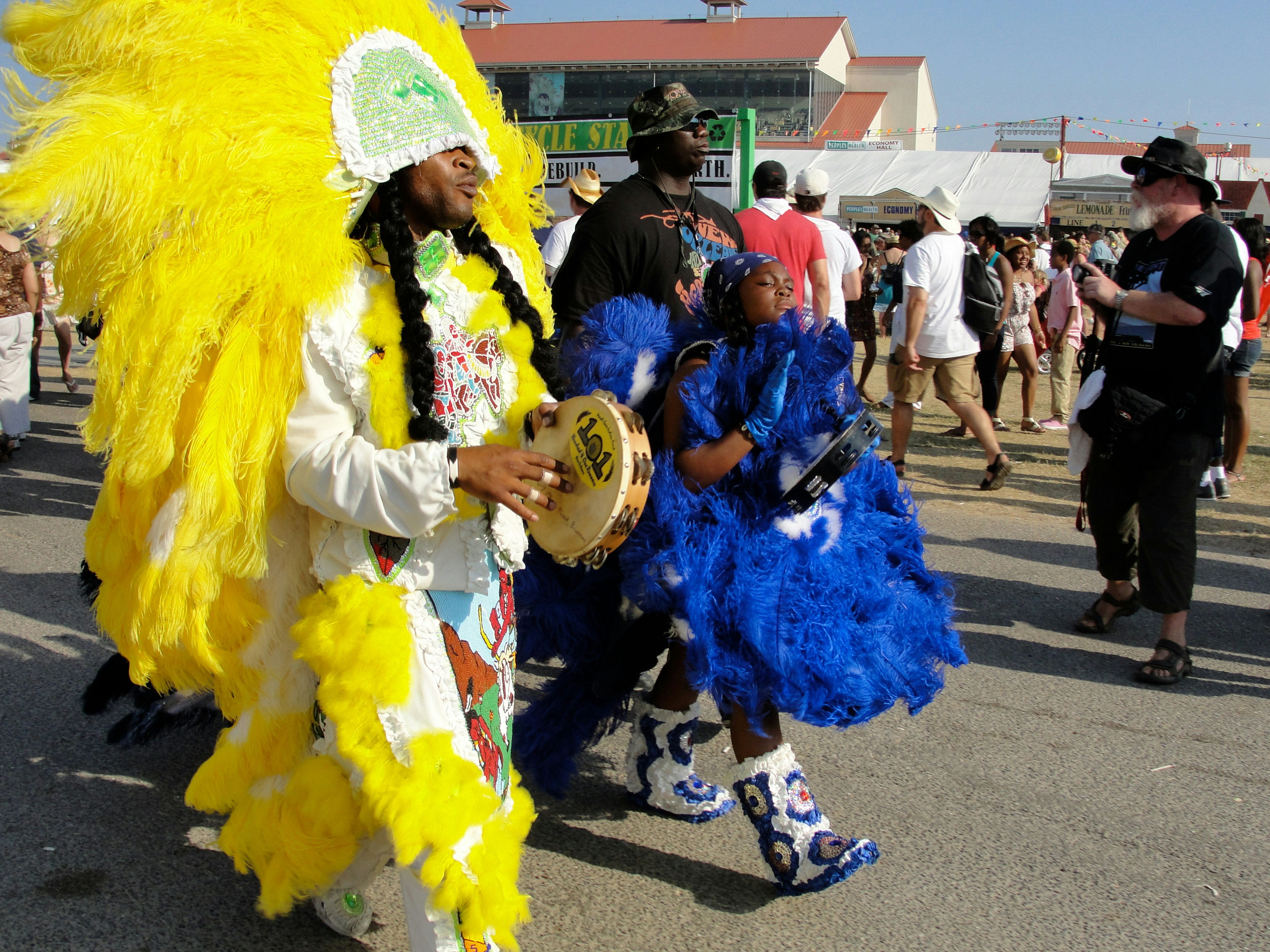 A man wearing a feathered head dress walks down a street carrying a tambourine