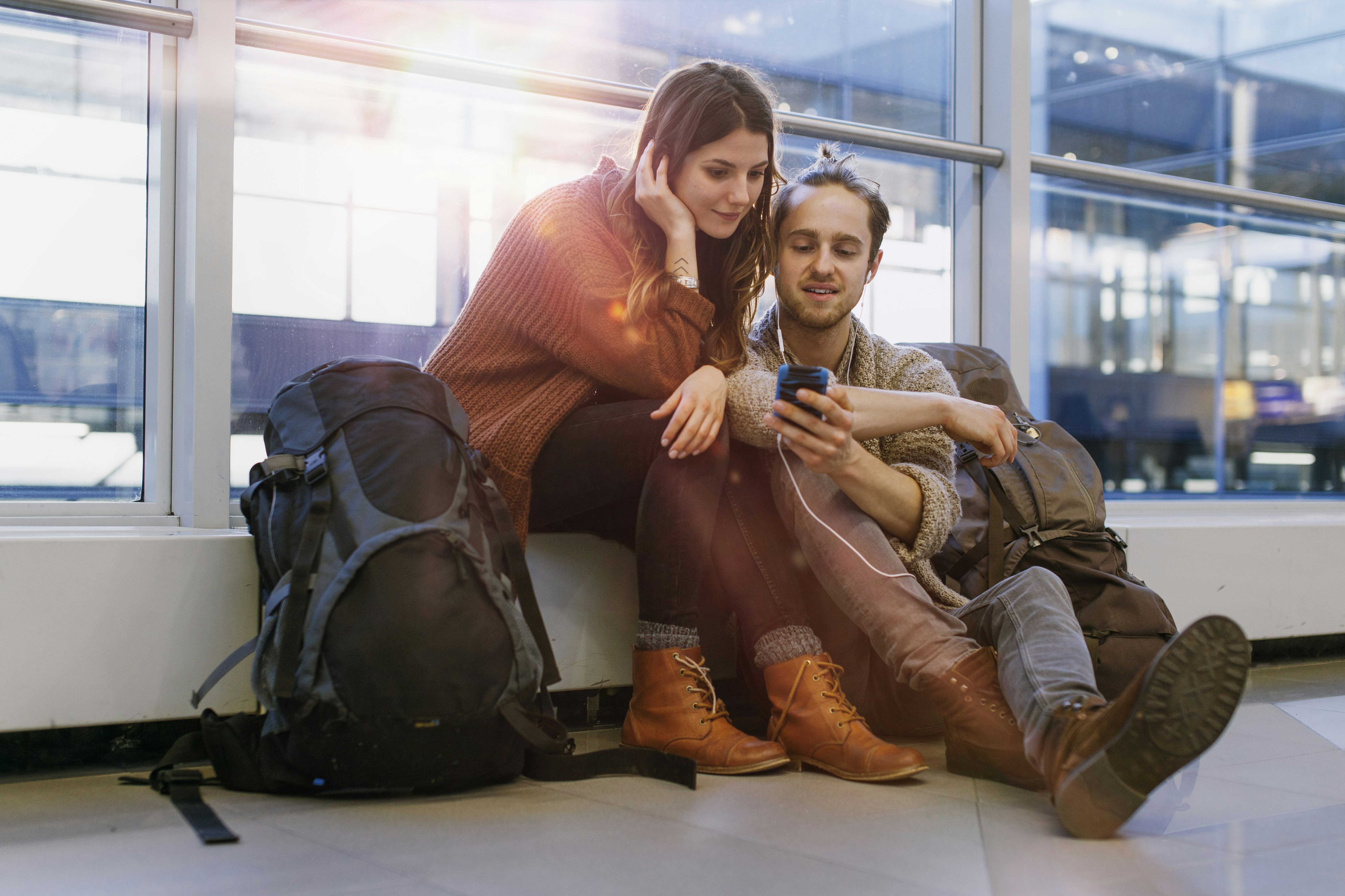 A young couple sitting on the floor in an airport terminal, passing time by watching something on a mobile phone. 