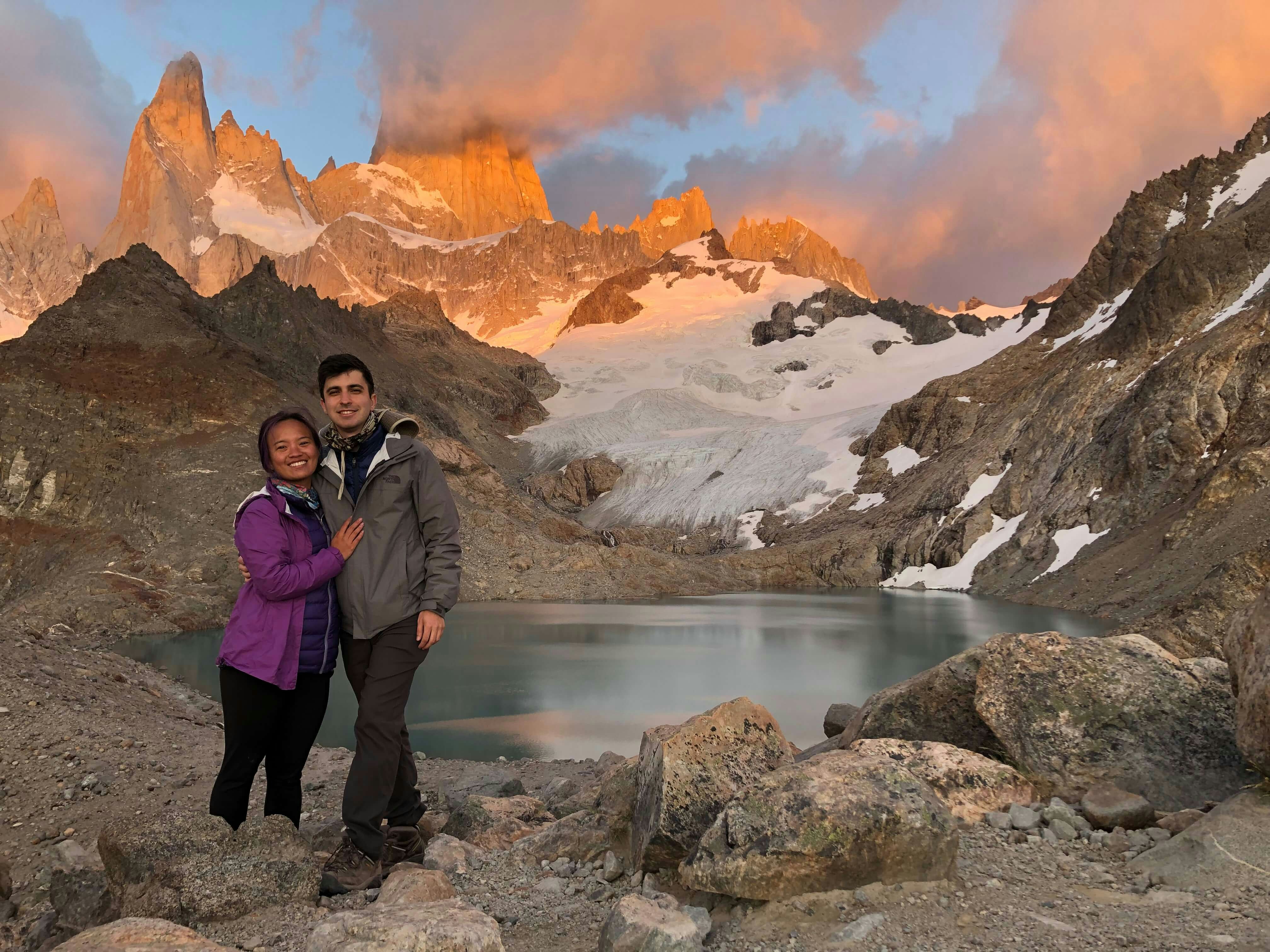 The young couple in hiking gear pose for a picture with a mountain and lake in the background at sunset.