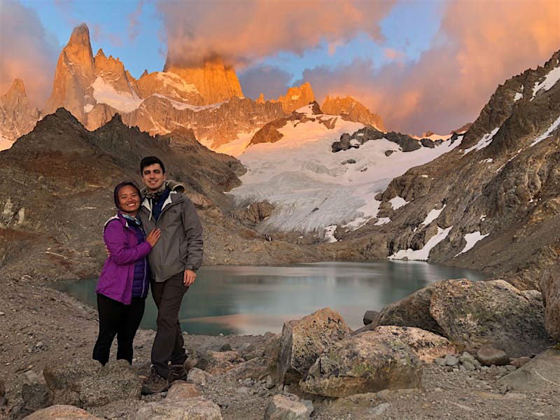 The young couple in hiking gear pose for a picture with a mountain and lake in the background at sunset.