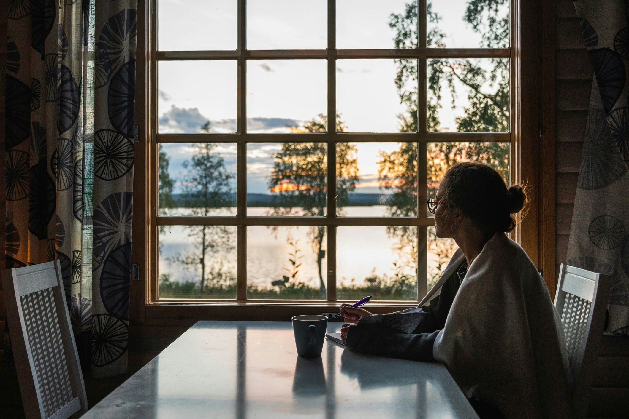 A young woman sits at a table with a cup of tea and a pen and a pad and looks wistfully out the window at a lake.