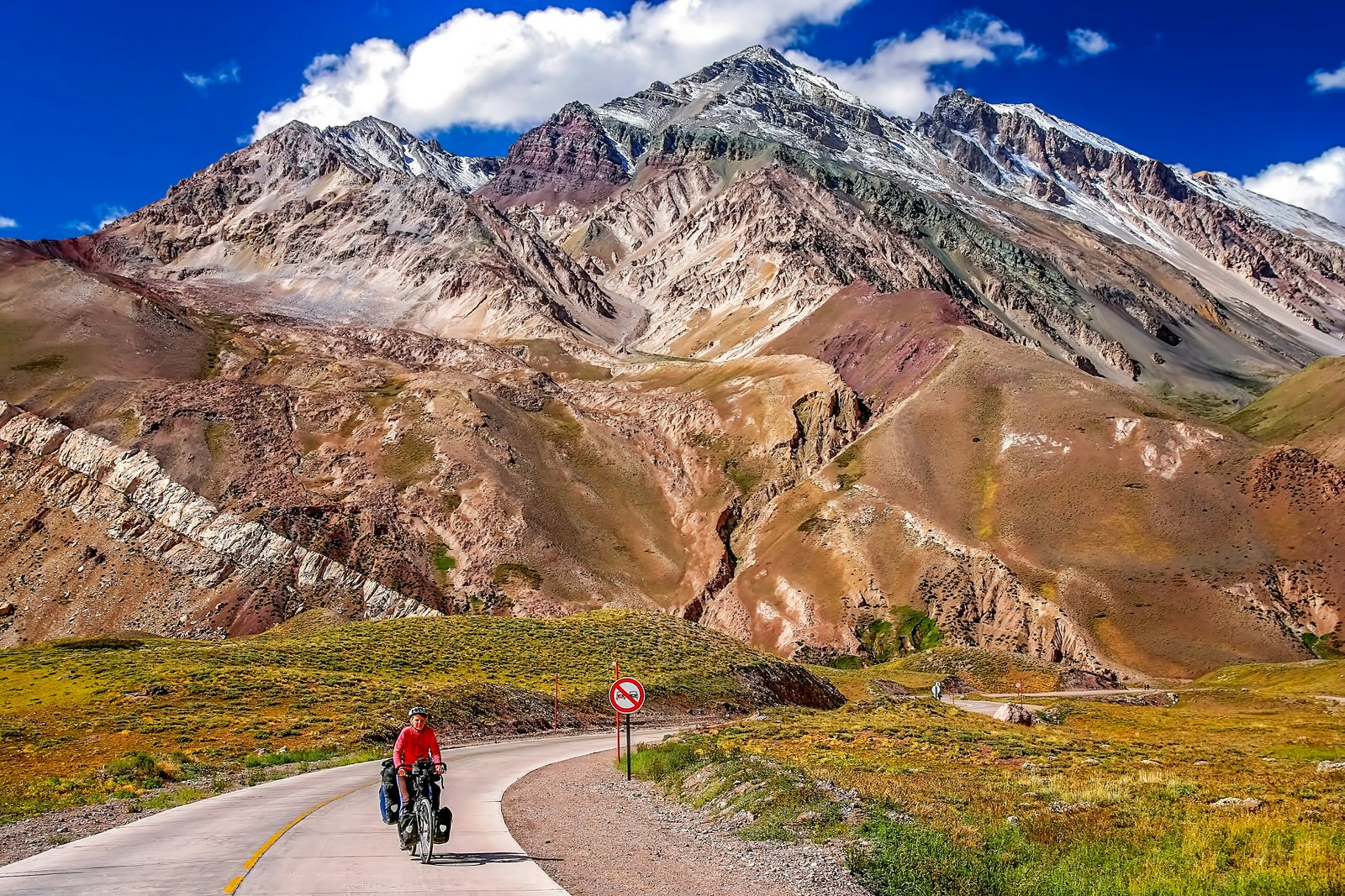 A cyclist, with bike laden with bags, cycles along a bending road beneath multi-coloured mountains.