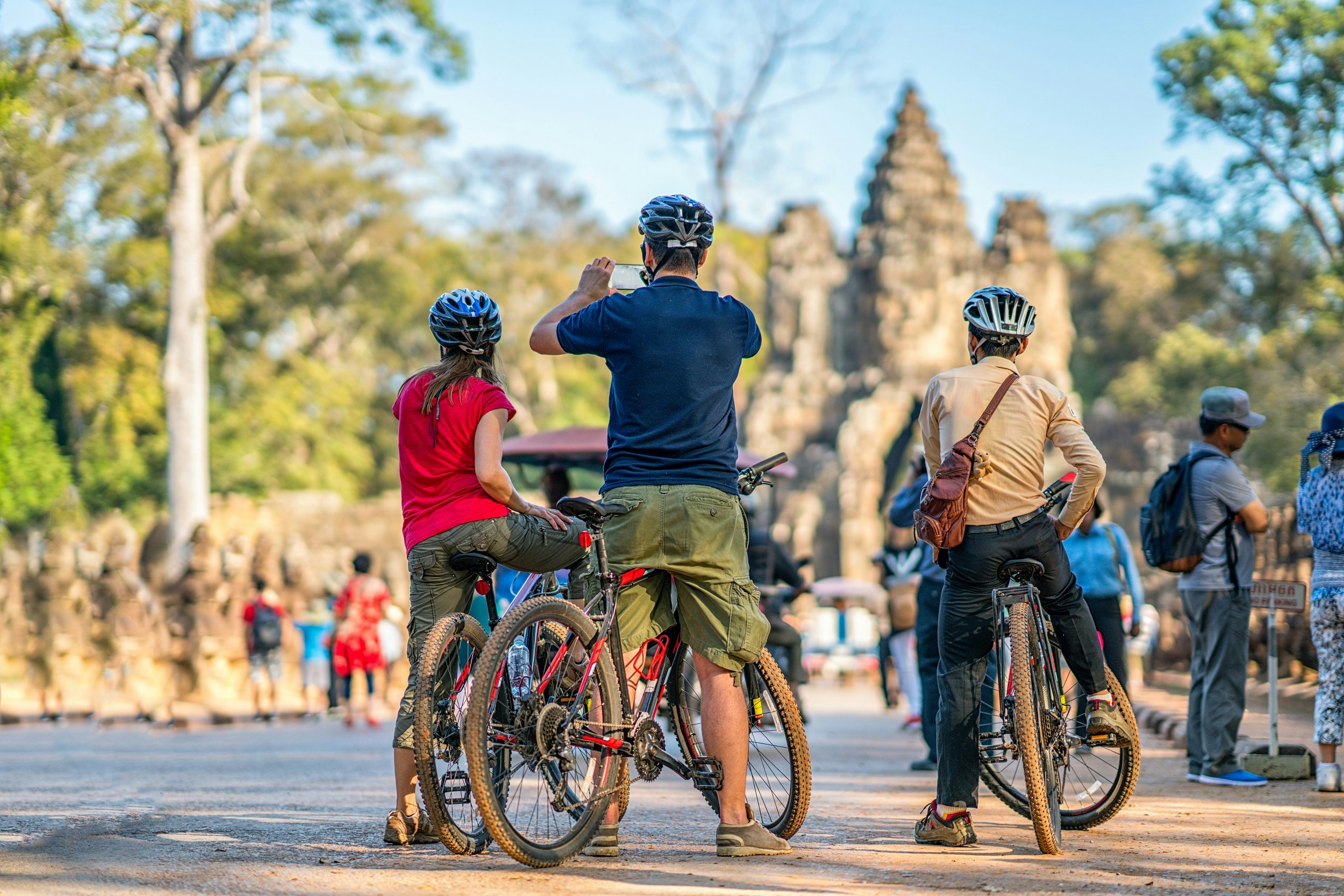 Three tourists, each wearing helmets, stand straddling mountain bikes; their backs are to the camera and they are facing some ancient ruins and crowds of people. The man in the middle is using his phone to take a picture.