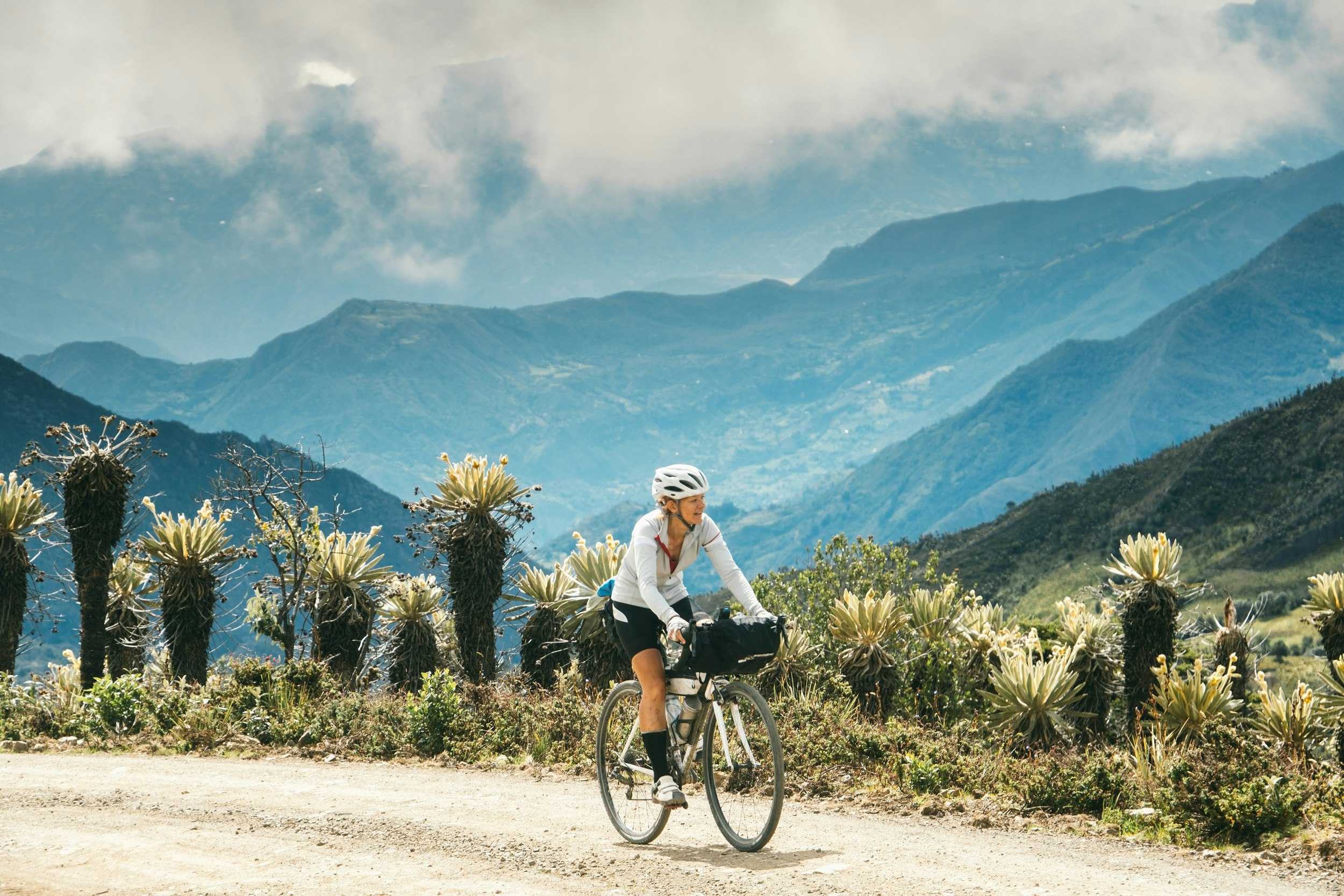 A woman cycles along a dirt road past large flowering cacti, with jungle covered mountain ridges in the distance.