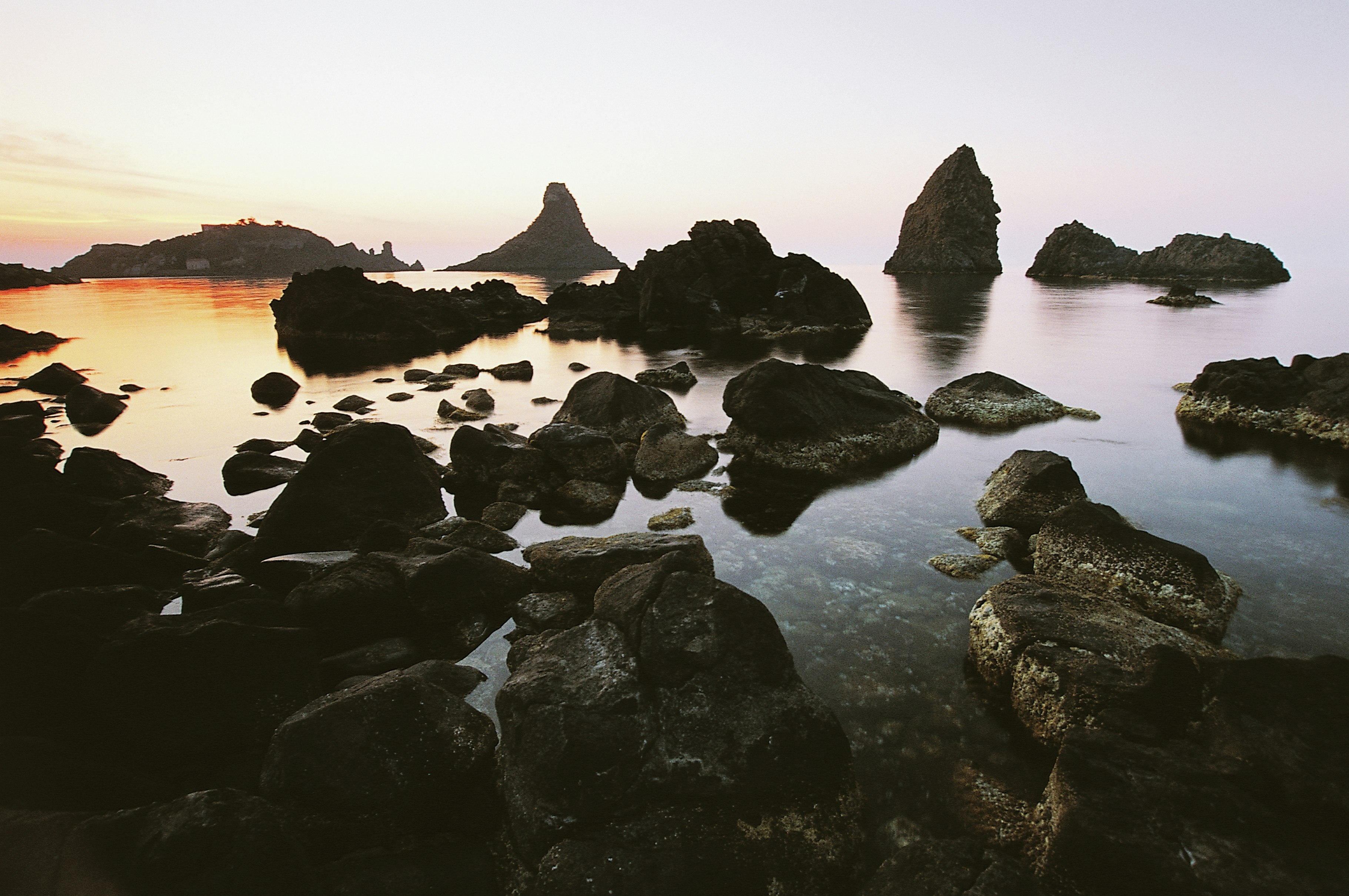 The sun sets creating a deep orange glow on the water on the left side of the frame contrasting with the cooler, pastel tones and dark grey of the basaltic rocks pointing out of the ocean