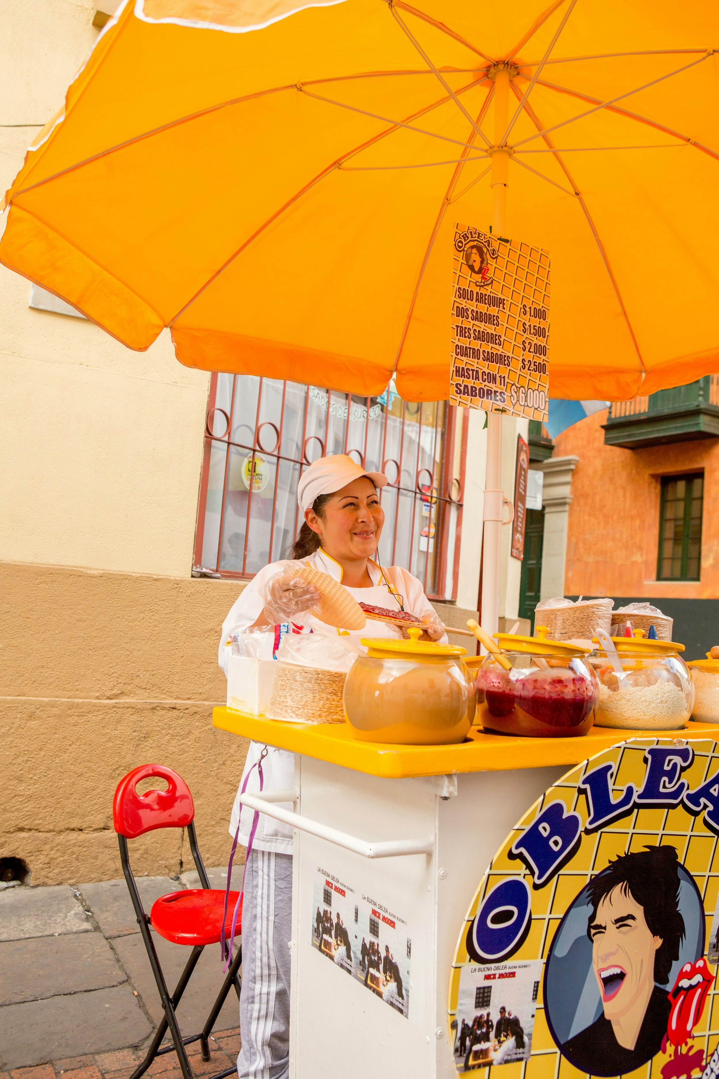 A smiling street vendor stands beneath a bright yellow umbrella that shelters her portable cart where she sells obleos; her sign has a cartoon of Mick Jagger and the Rolling Stones logo.