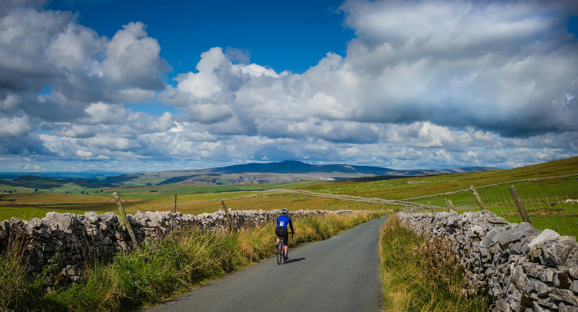 A lone cyclist pedals along a narrow stretch of road lined by old stone walls; the road meanders into the distant hills.