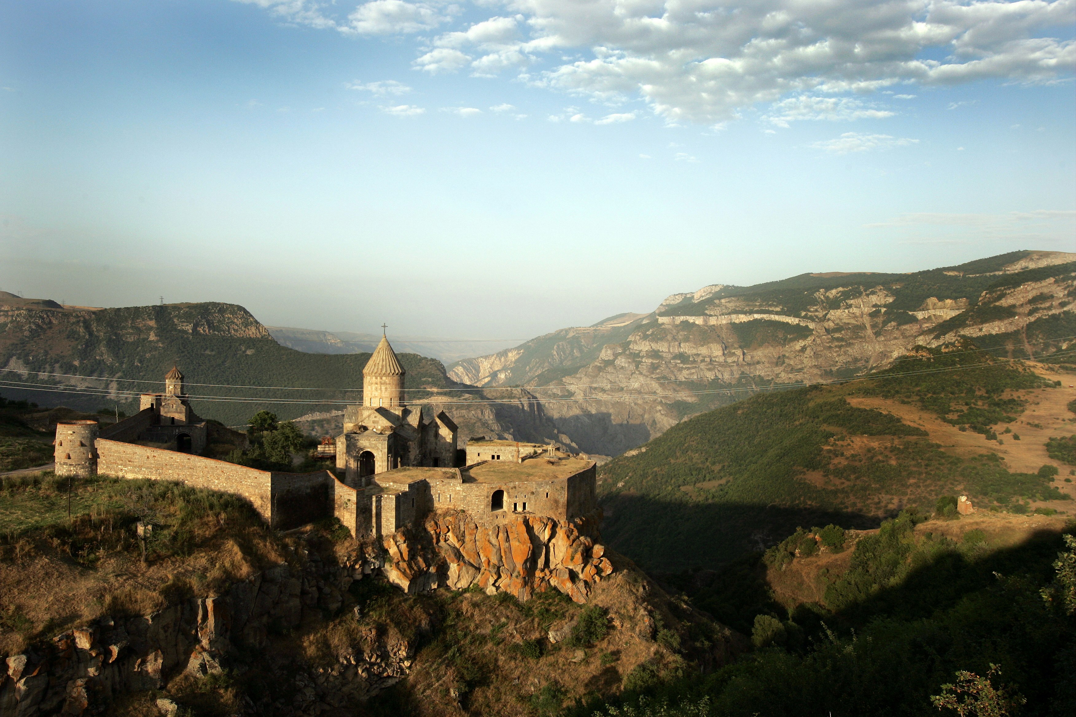 The monastery at Dadivank is situated on a rocky ridge with some green scrub grass covering the orange volcanic grown. In the background are more rocky ridges and hills extending to a cloud-lined horizon.