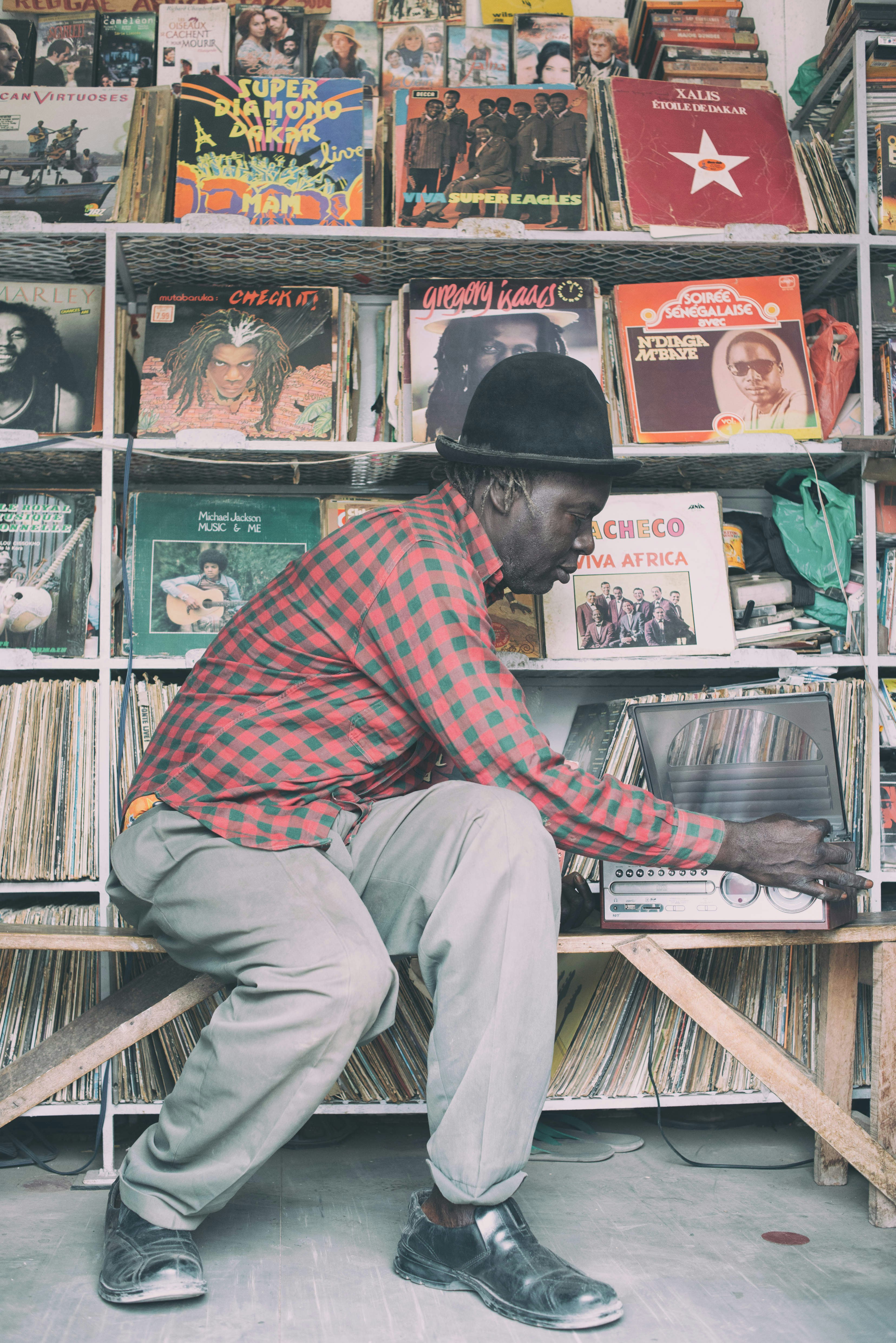 Dread Amala leans over his record player, adjusting the contols; behind him are shelves upon shelves of classic albums.