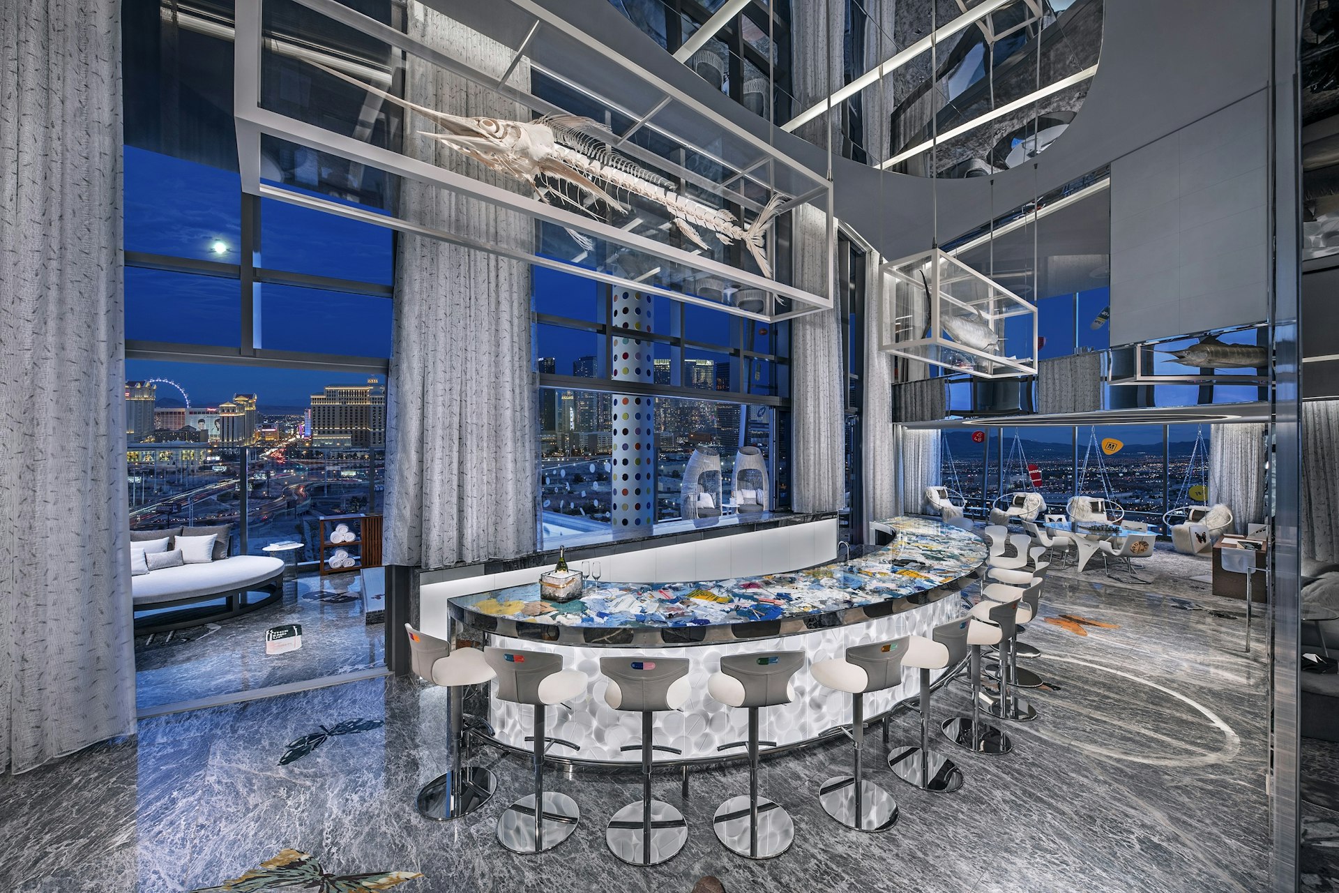 A large apartment space with floor-to-ceiling glass windows allowing a view of Las Vegas. The room is grey and white with splashes of colour, often in the shape of butterflies. A bar forms a half circle in the centre of the room, with ten empty stools around it