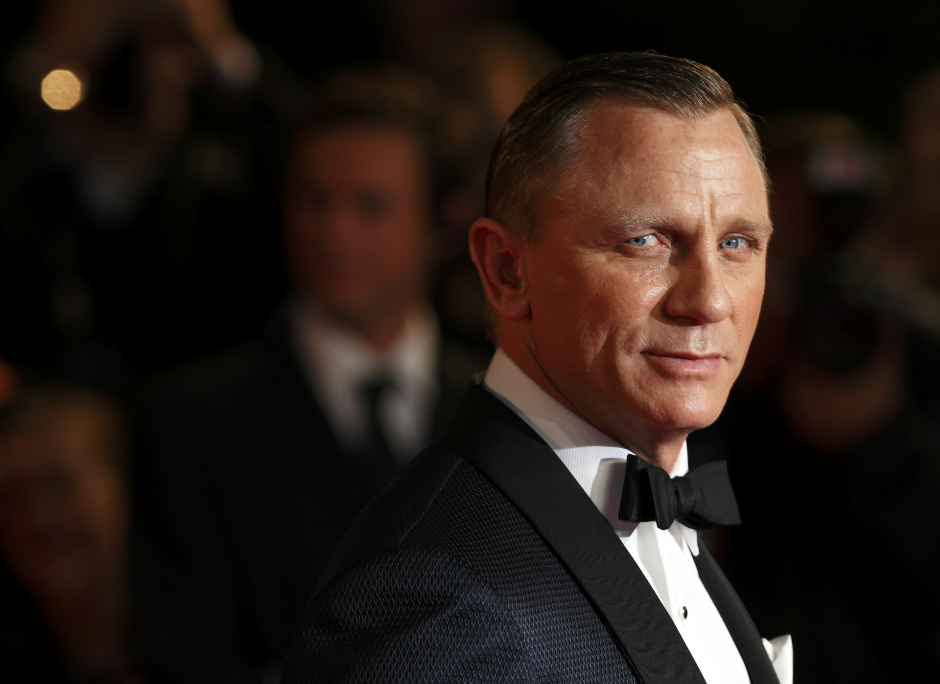 James Bond actor Daniel Craig is wearing a tuxedo and looking towards the camera; the background is dark and in soft focus.