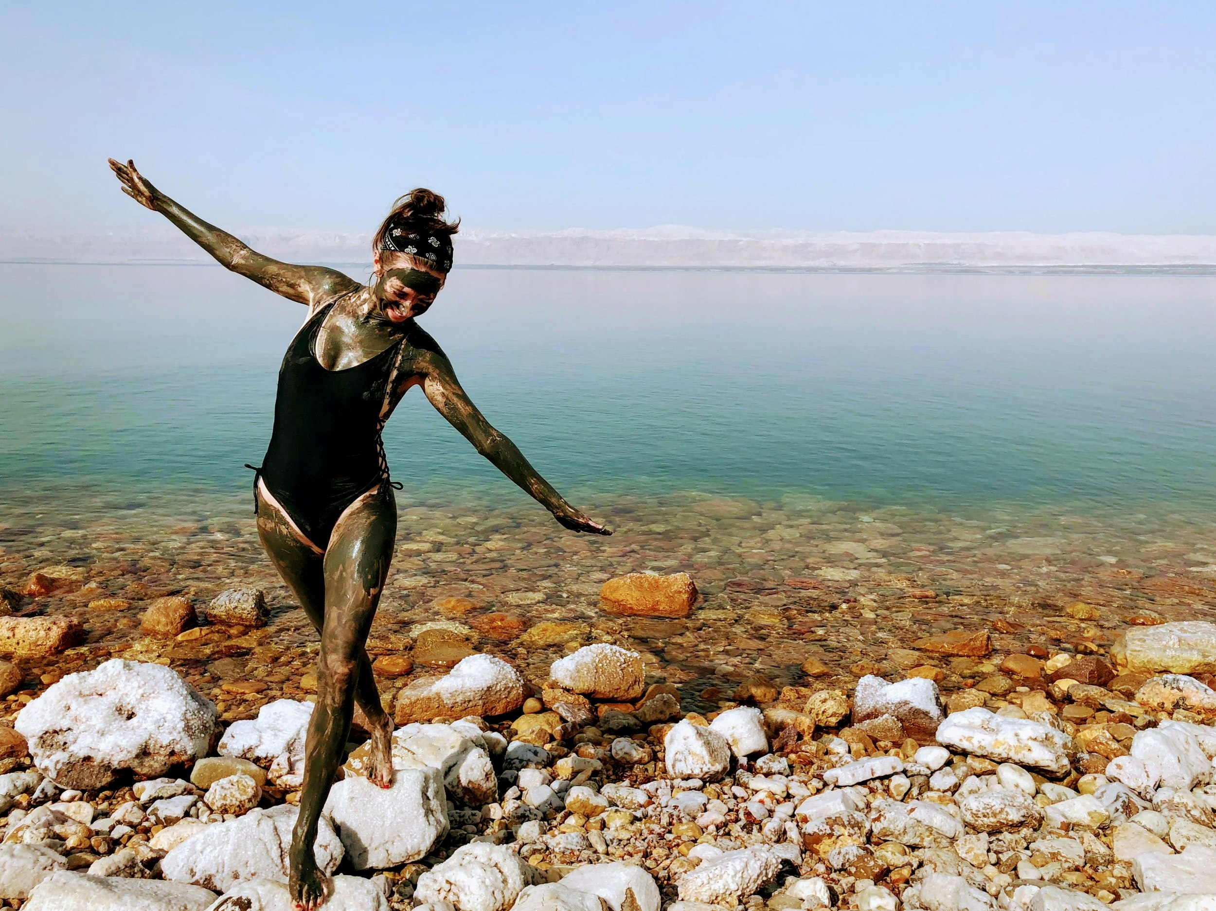 A woman in a black bathing suit stands on some white rocks at the shore of the Dead Sea; her arms are spread wide and her legs are crossed, while her legs, arms and face is covered in black mud.