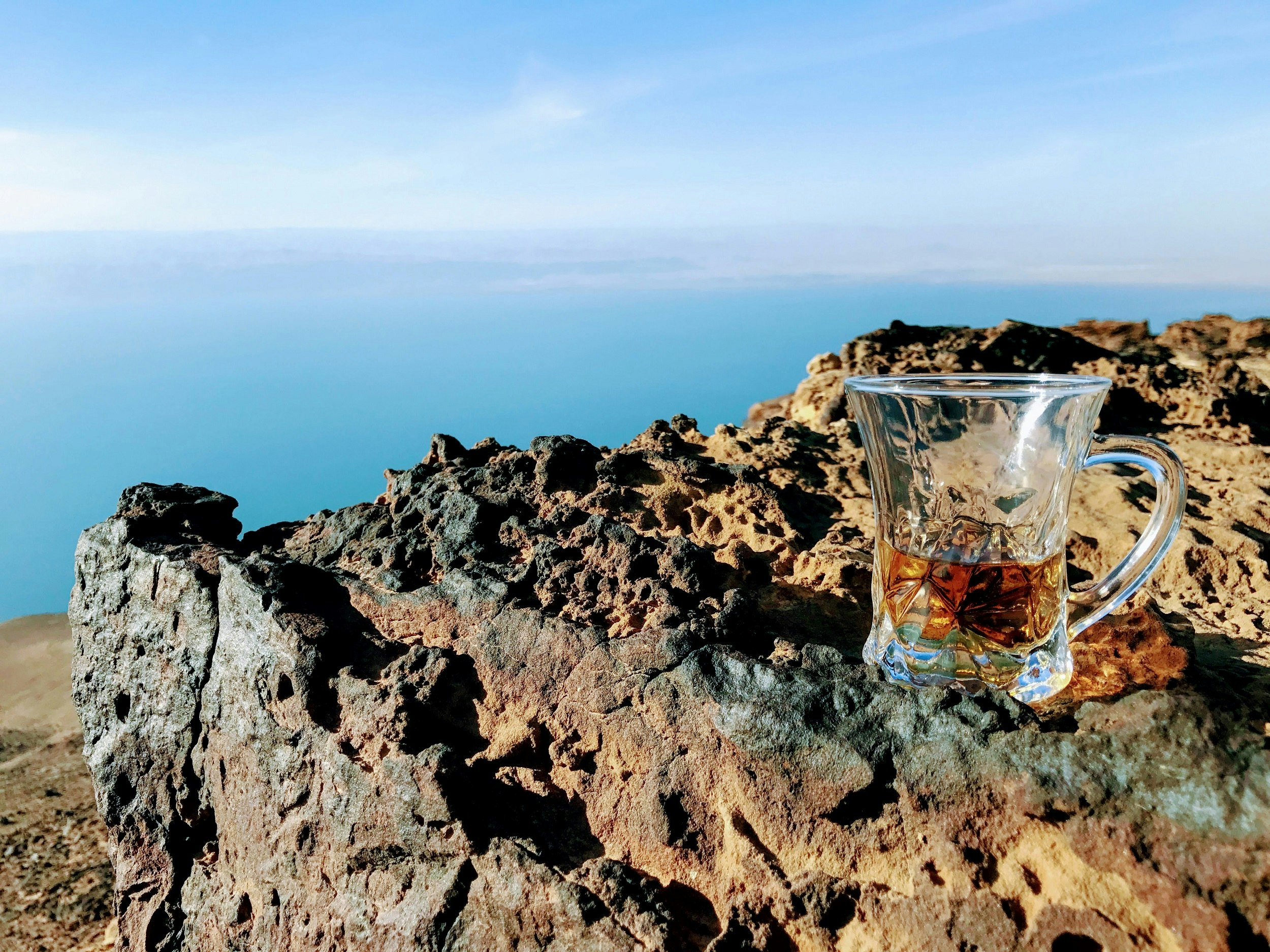 A decorative glass cup (half-full of tea) with handle sits on a rust-coloured rock overlooking the Dead Sea.