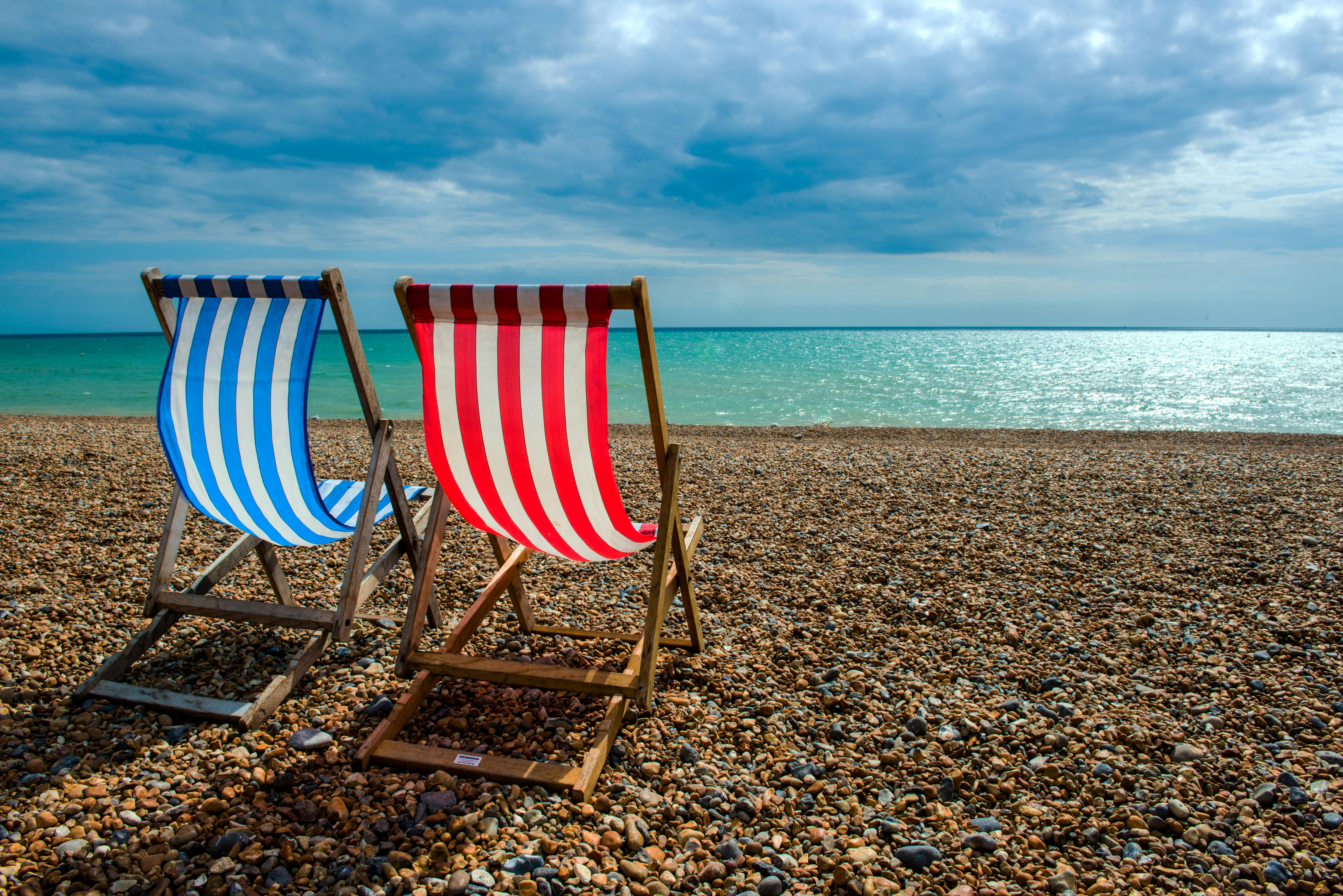 Two empty deck chairs, one with red and white stripes, the other with blue and white stripes, are arranged to look out towards the sea on Brighton beach. The fabric is billowing in the wind.