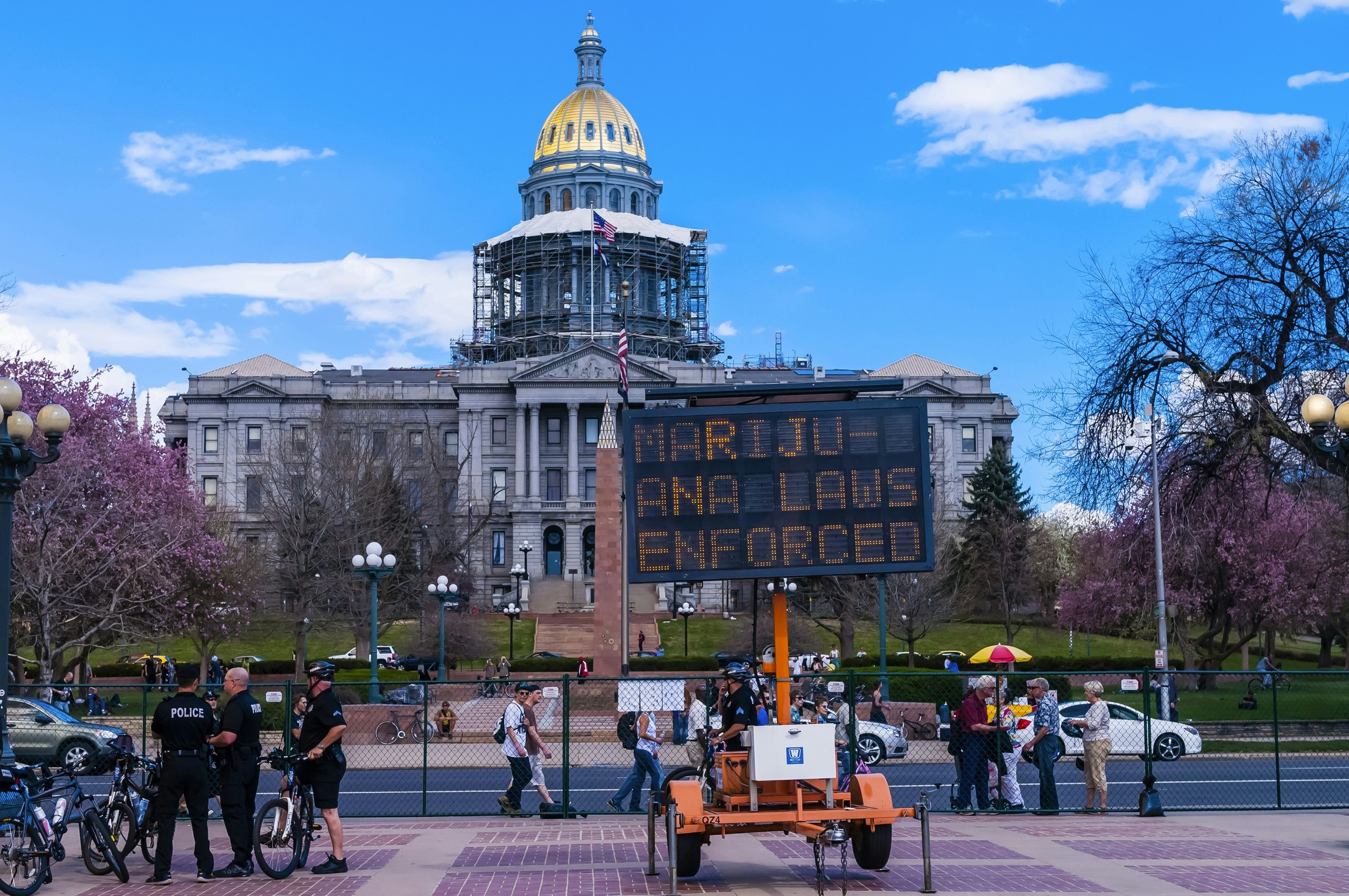 Three bicycle cops stand near an electronic billboard reading "Marijuana Laws Enforced" in front of the Colorado State Capitol Building in Civic Center Park in Denver. The trees are covered in purple spring blooms that mirror the reddish color of the paved sidewalk. 