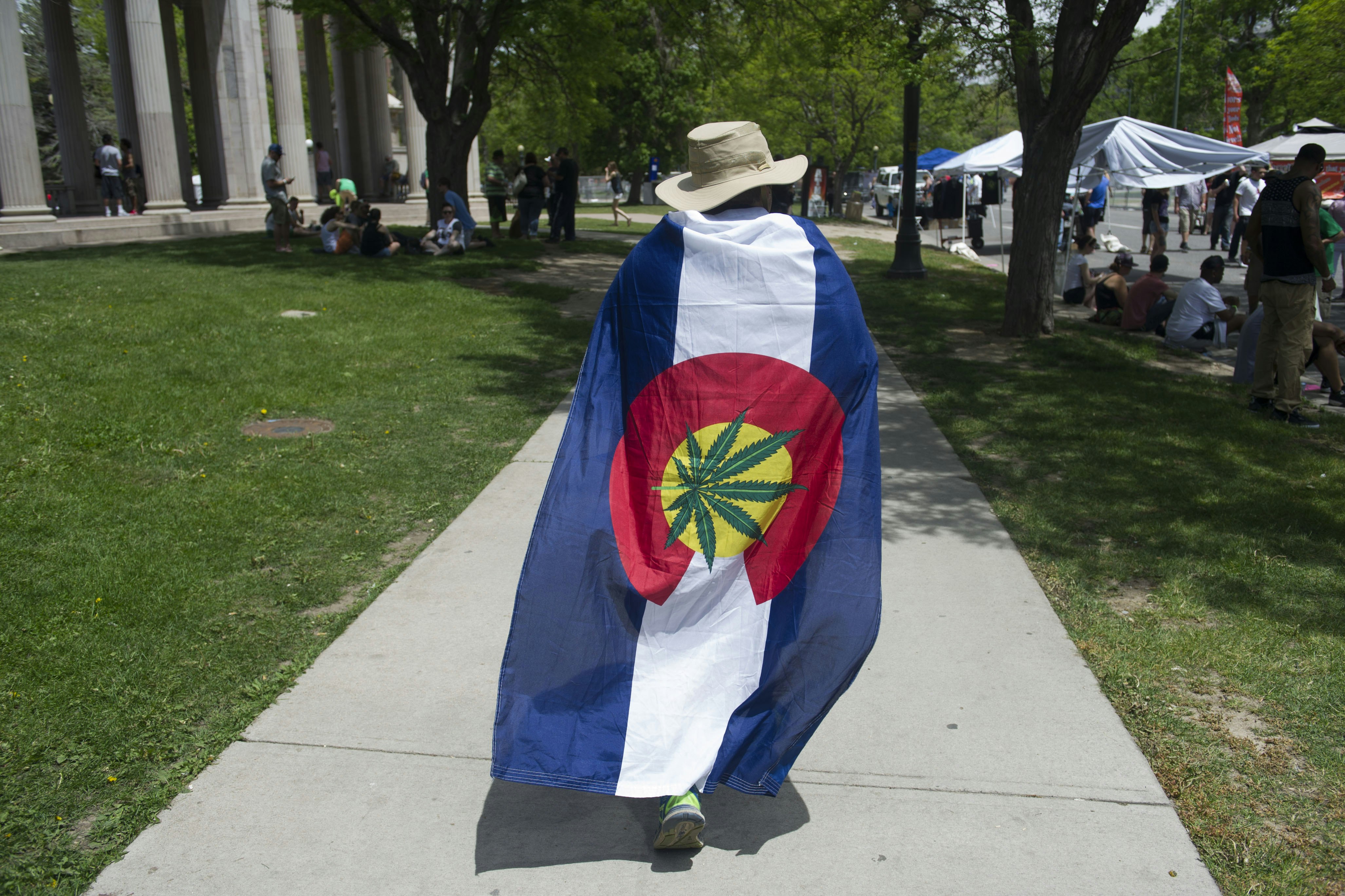 A man in a white bucket hat walks down the sidewalk wearing a Colorado state flag as a cape, where the yellow center of the flag is overlaid with a green cannabis leaf
