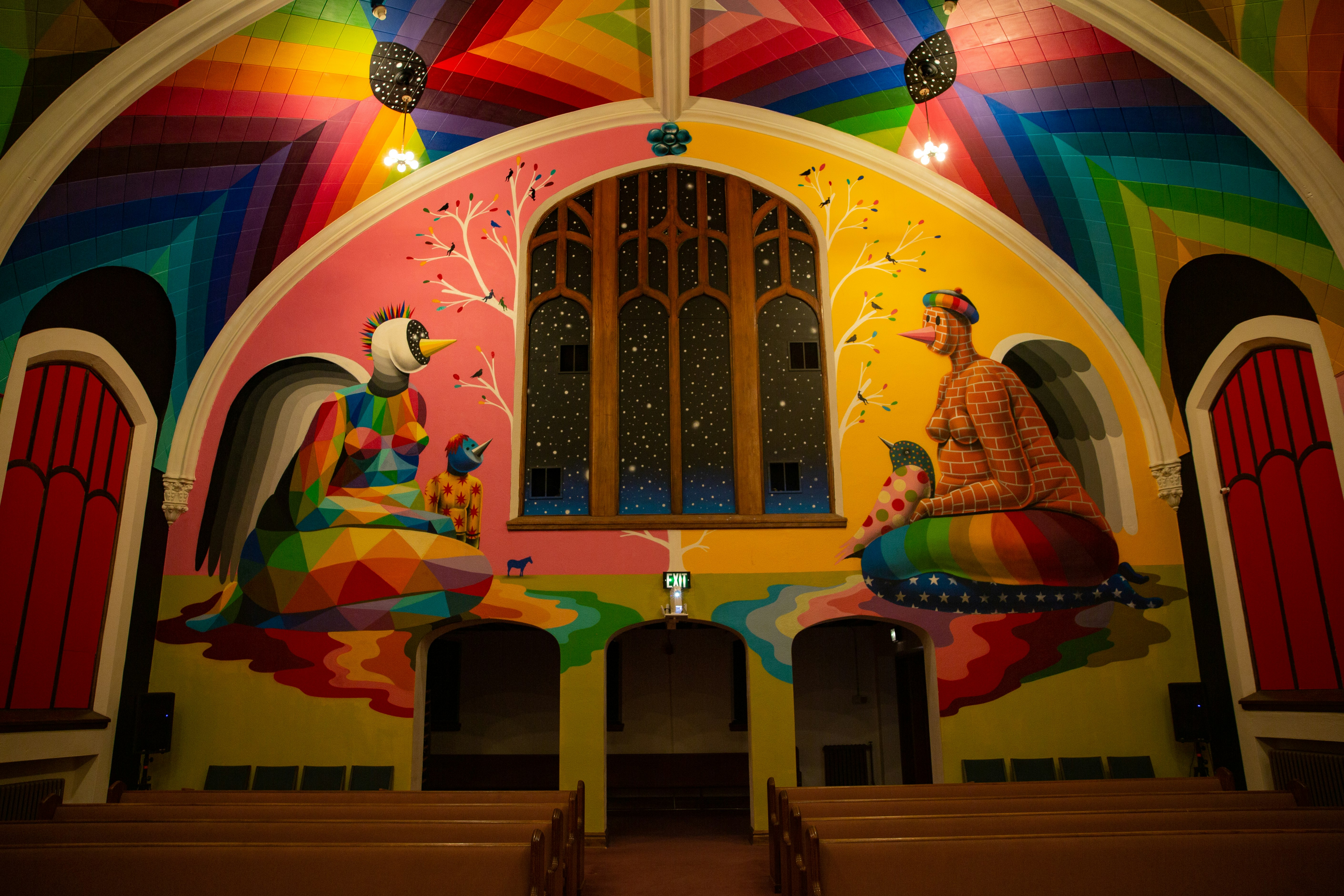 The colorful interior of the Denver Church of Cannabis is made up of a vaulted ceiling painted in a mural of rainbow hues portraying two kneeling female figures with bird-like beaks, one painted with geometric shapes and the other with a brick pattern. A three-paned arch window has warm wood trim and beneath are three arched doors in front of rows of wooden pews.