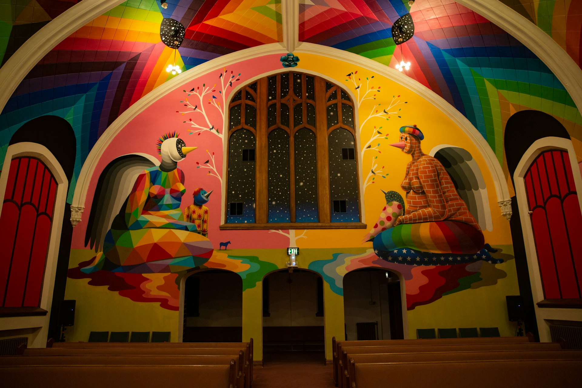 The colorful interior of the Denver Church of Cannabis is made up of a vaulted ceiling painted in a mural of rainbow hues portraying two kneeling female figures with bird-like beaks, one painted with geometric shapes and the other with a brick pattern. A three-paned arch window has warm wood trim and beneath are three arched doors in front of rows of wooden pews.