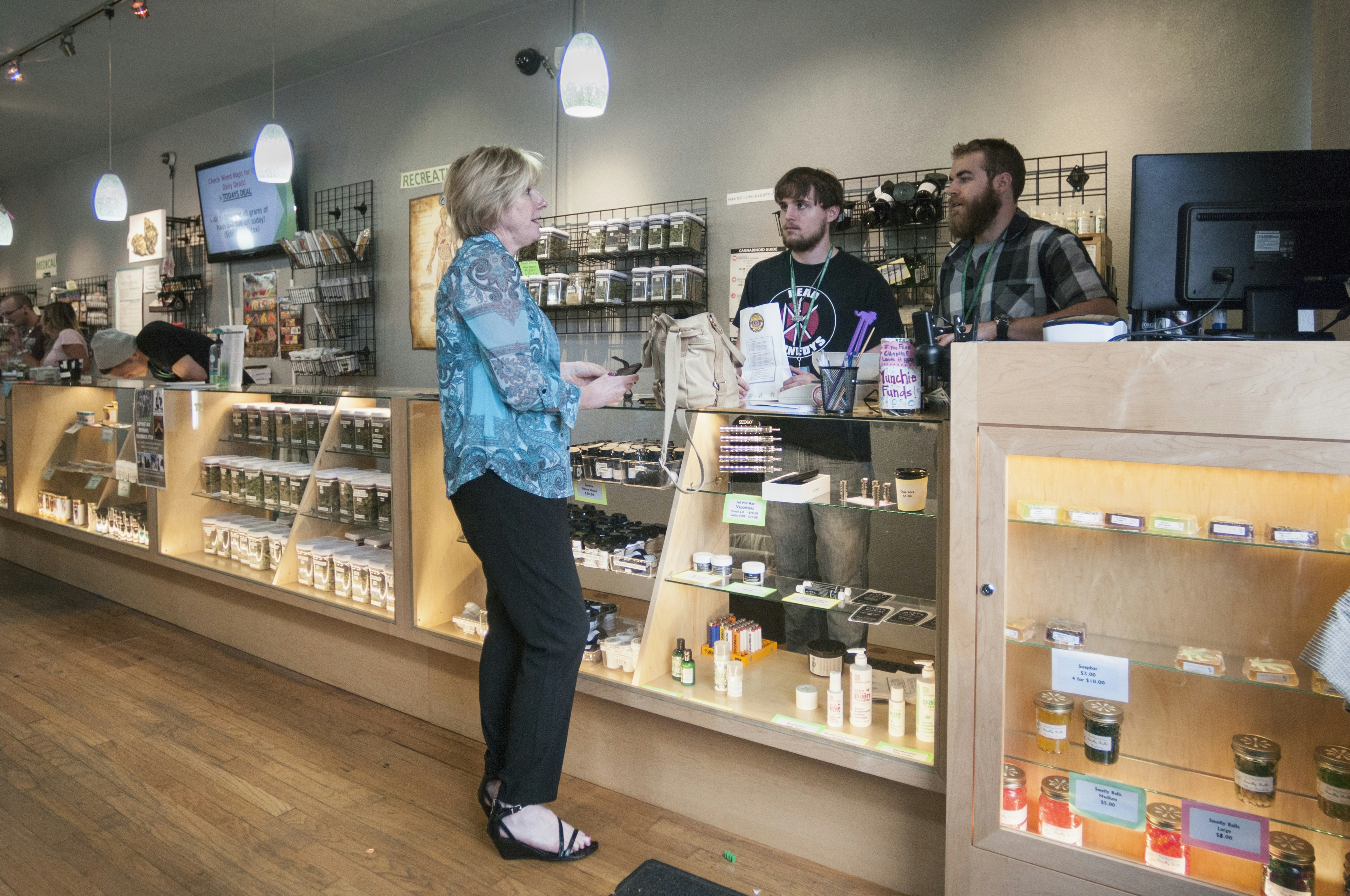 A middle aged white woman with short blond hair in a blue floral tunic, black slacks, and black sandals speaks with two budtenders at a Denver, Colorado cannabis dispensary. A long display case and counter made of blonde wood and glass shows cases a variety of edible and topical cannabis products and concentrates, while jars of flower sit on shelves on the wall