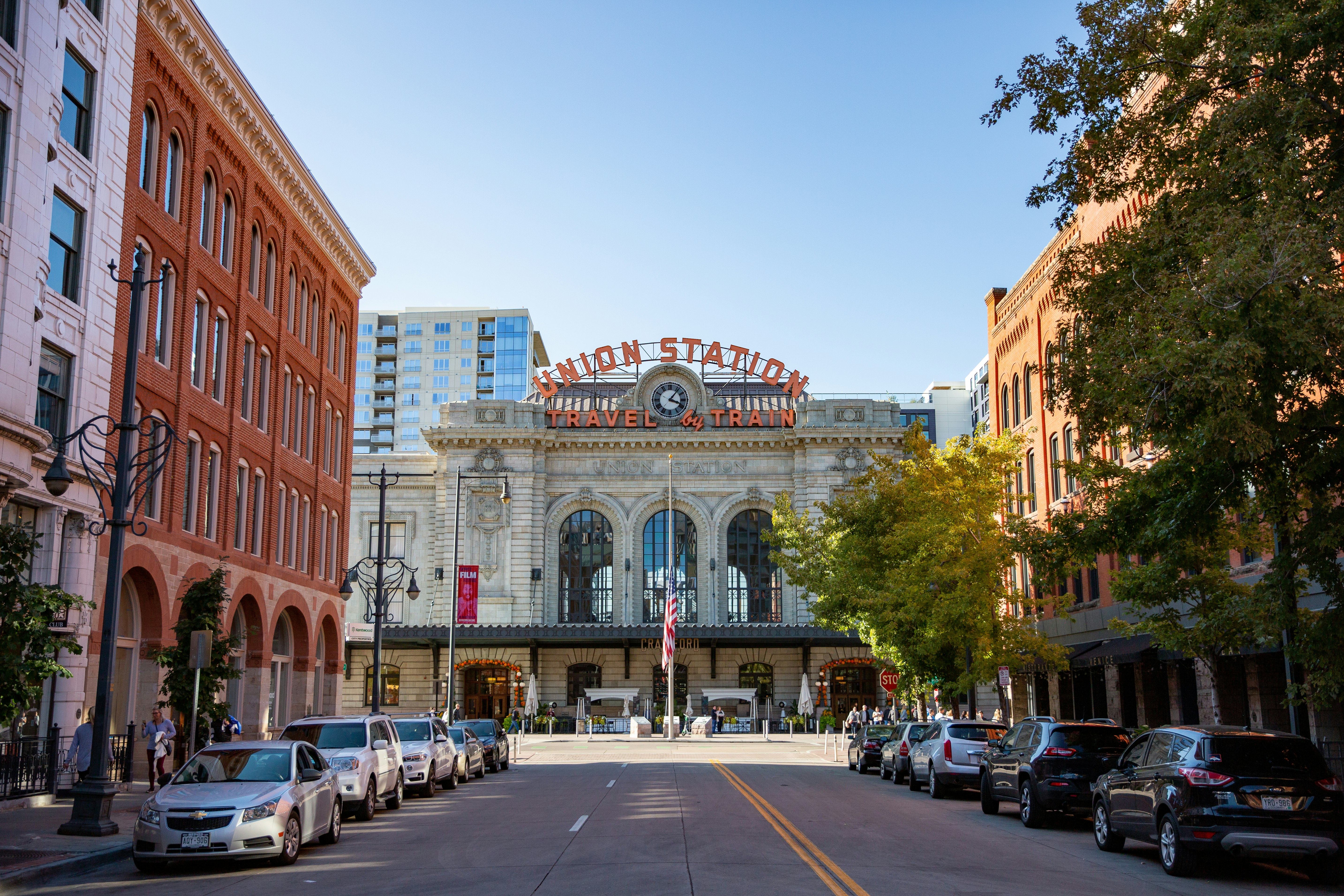 Brick buildings line a black paved street that dead ends in the stone facade of Union Station, which also features a white clock face and a bright red sign that reads "Union Station Travel By Train" 