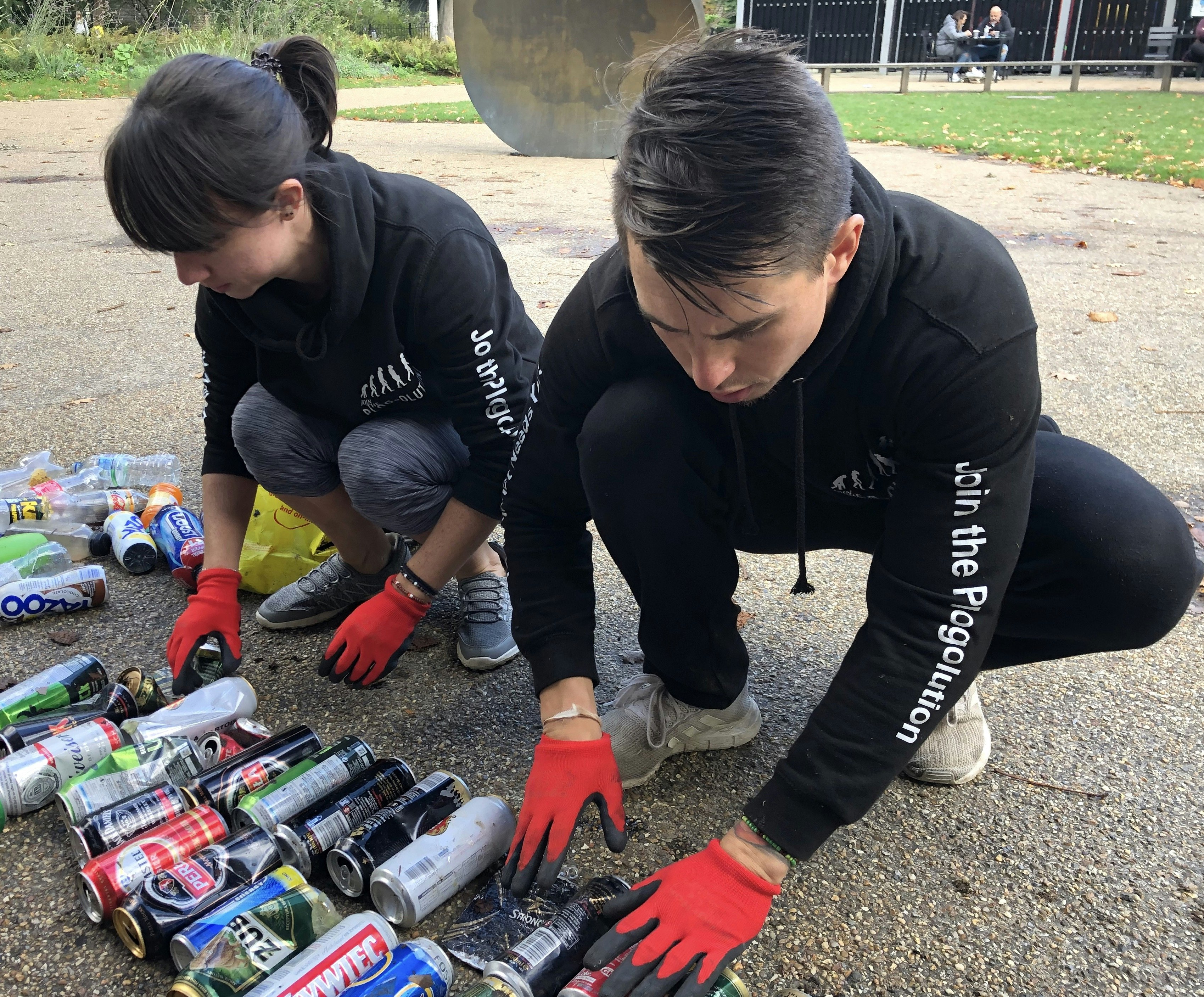 Dermot Kavanagh and Phoebe Abrahams sort through discarded cans of beer, while crouching on the ground. Both are dressed in all-black jogging gear emblazoned with the slogan 'join the plogolution'.