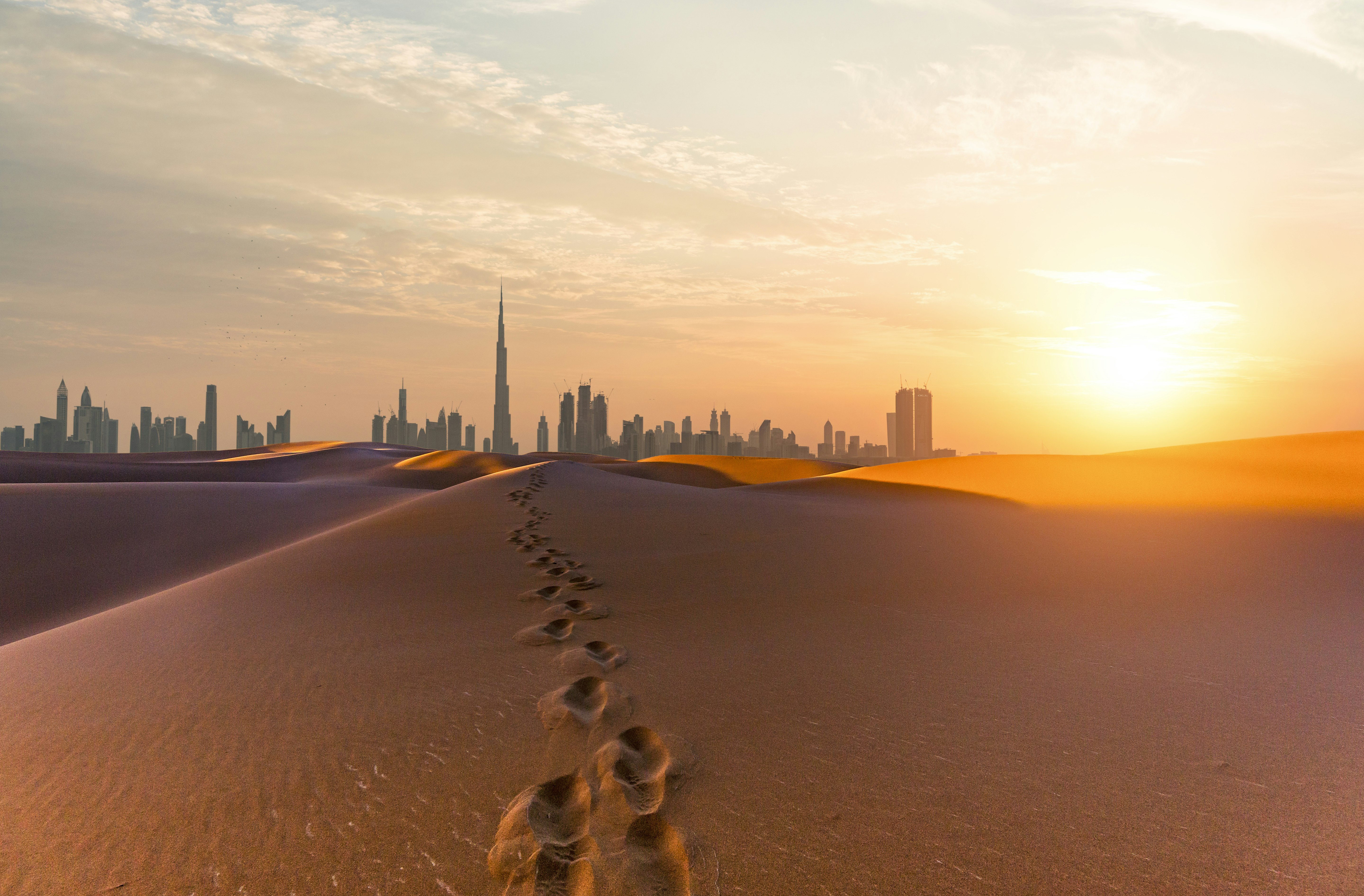Footsteps in the desert sand heading towards the skyscrapers of the Dubai city skyline at dawn.