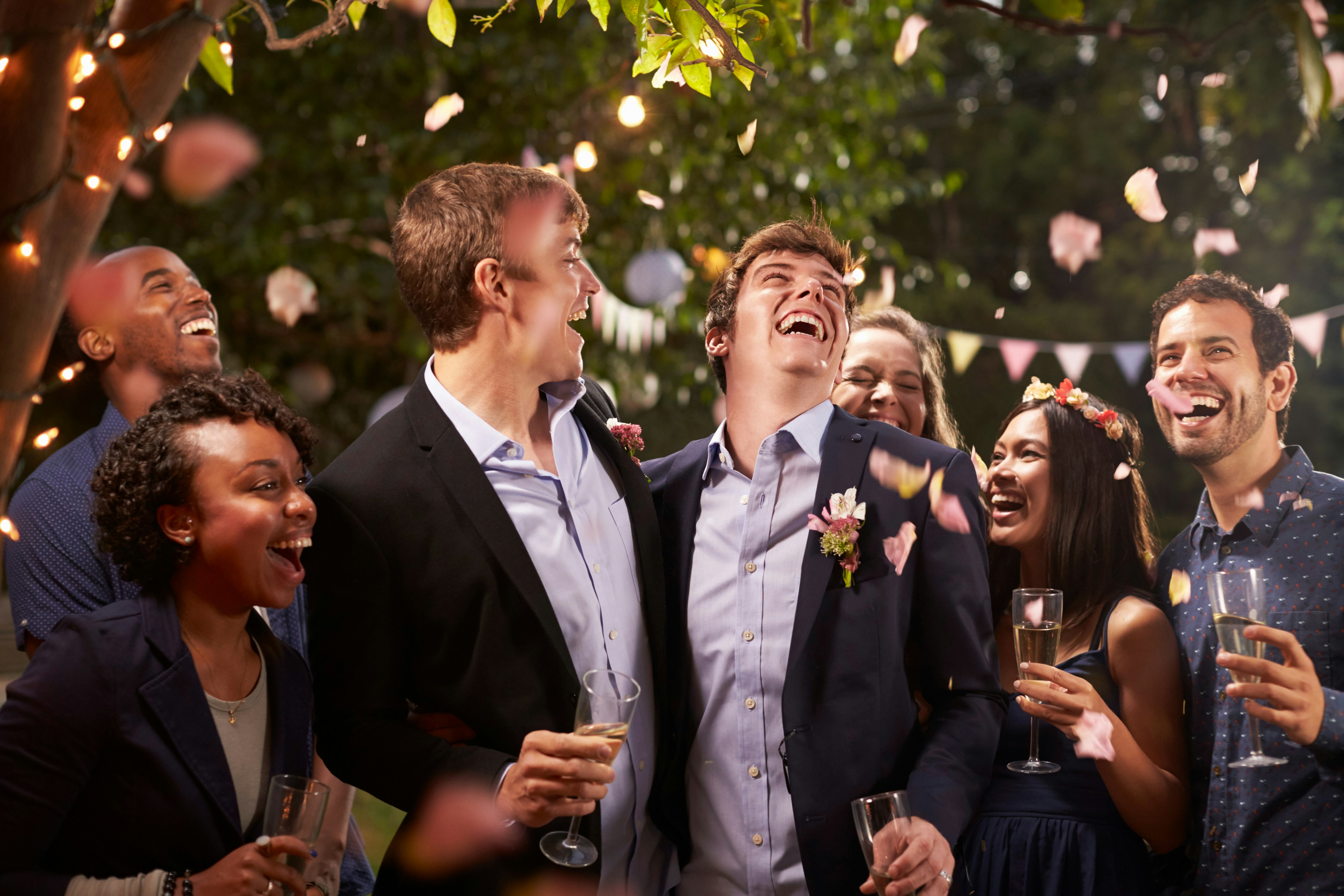 Two men standing under confetti, celebrating their marriage at a wedding reception surrounded by loved ones.