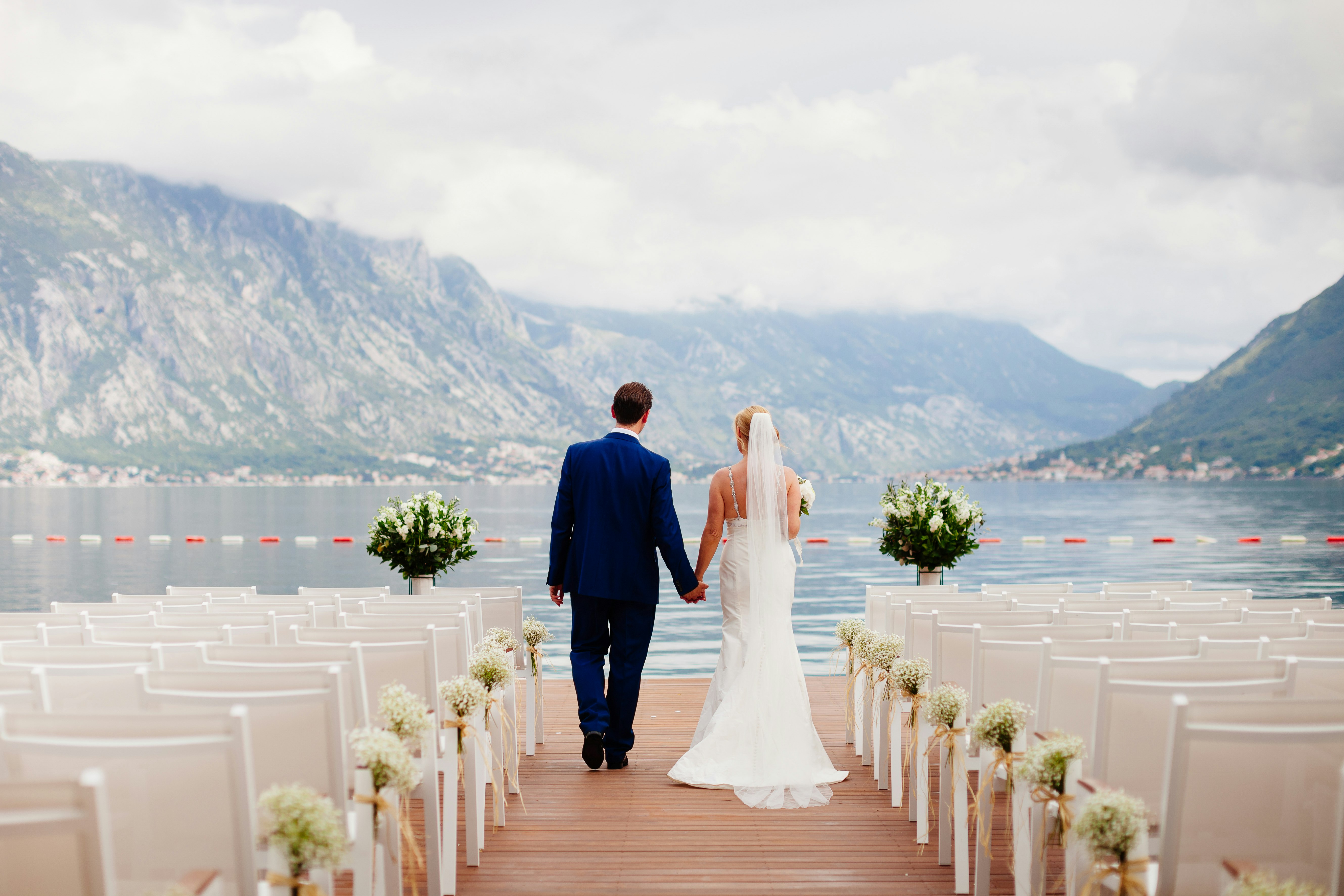 A bride and groom walk between sets of empty chairs laid out for a wedding ceremony in a mountainous lakeside setting.