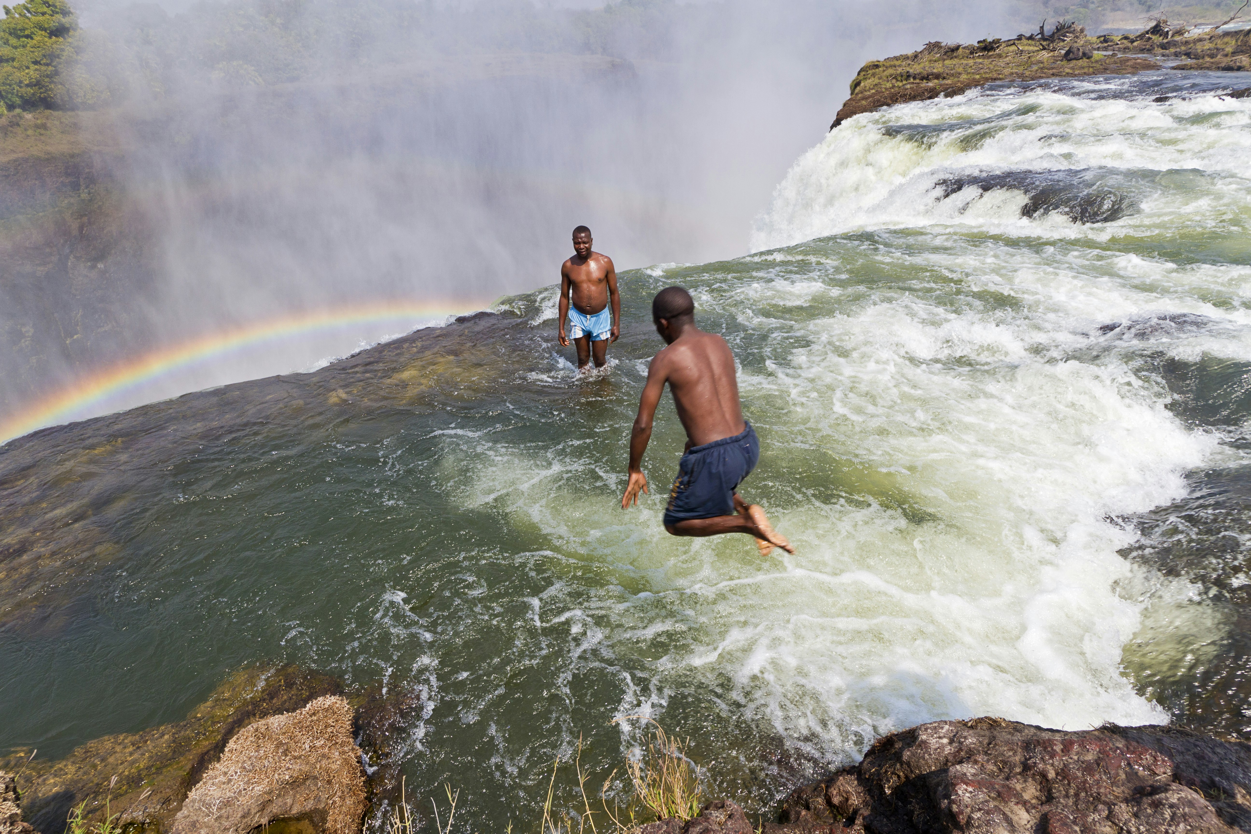 Two men in swim trunks enjoy Devil's Pool atop Victoria Falls, one standing close to the edge watching as the other jumps from a high point into the pool below with his back to the viewer