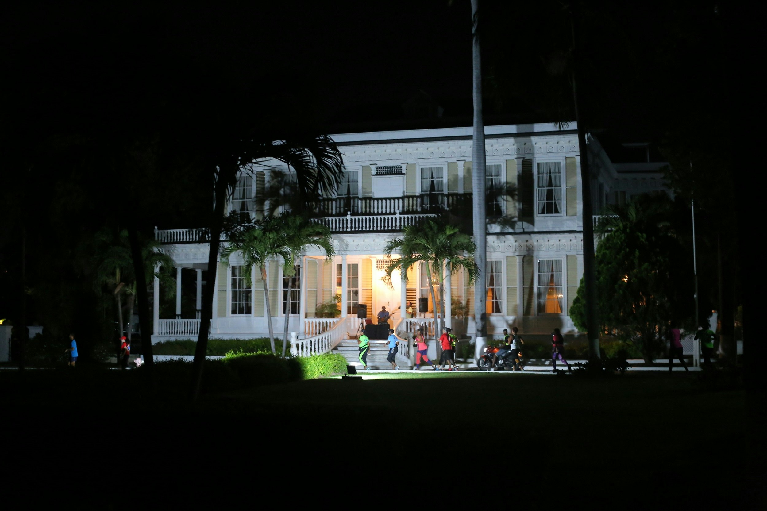 Runners make their way in front of a historic mansion; it's dark outside, but the house is floodlit, and there is a musician playing on the verandah.