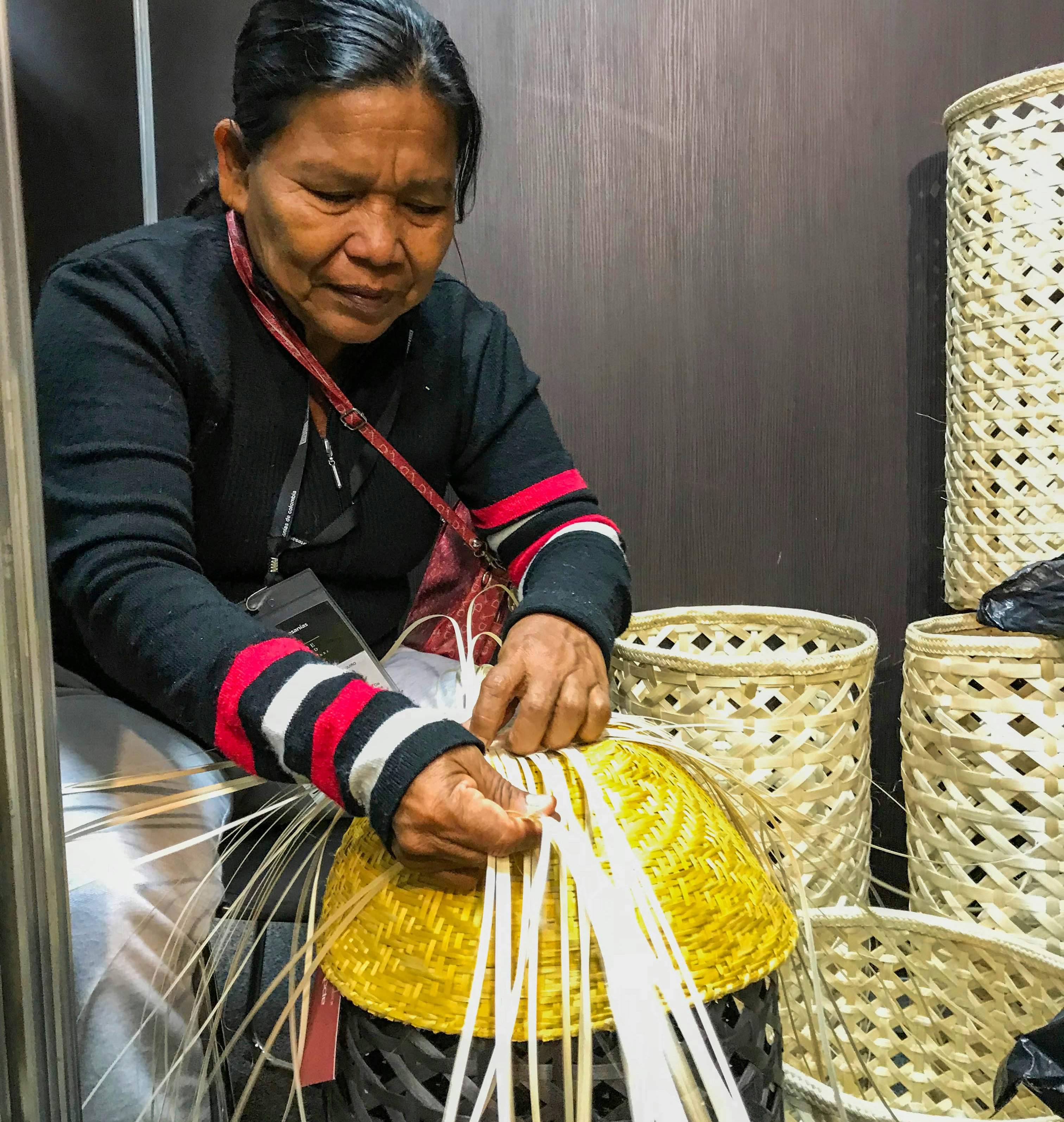 A woman with black hair and a black sweater with red and white stripes on the cuffs works on weaving a basket out of several flat strands of a pale material. To the right of her and the basket she is working on are several finished pieces, tall cylindrical baskets with a diamond pattern to them