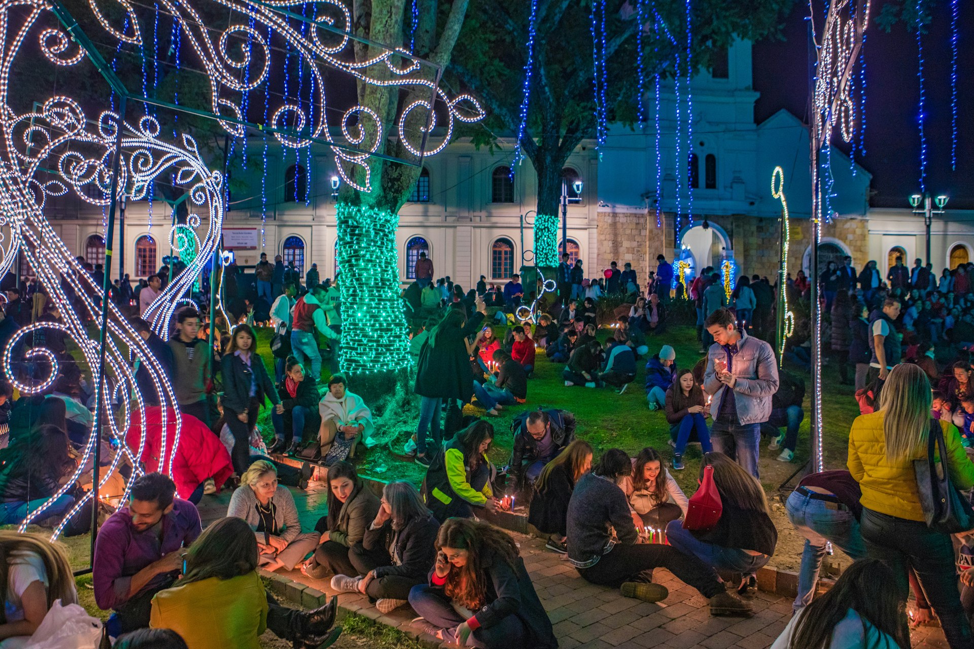 Crowds of people celebrate Dia de Las Velitas by lighting candles and enjoying green Christmas lights wrapped around tree trunks, strands of blue lights dangling from the trees like moss, and white light sculptures in the shape of curlicues and vines standing upright by the sidewalk. In the background are tall white colonial-era stone buildings. 