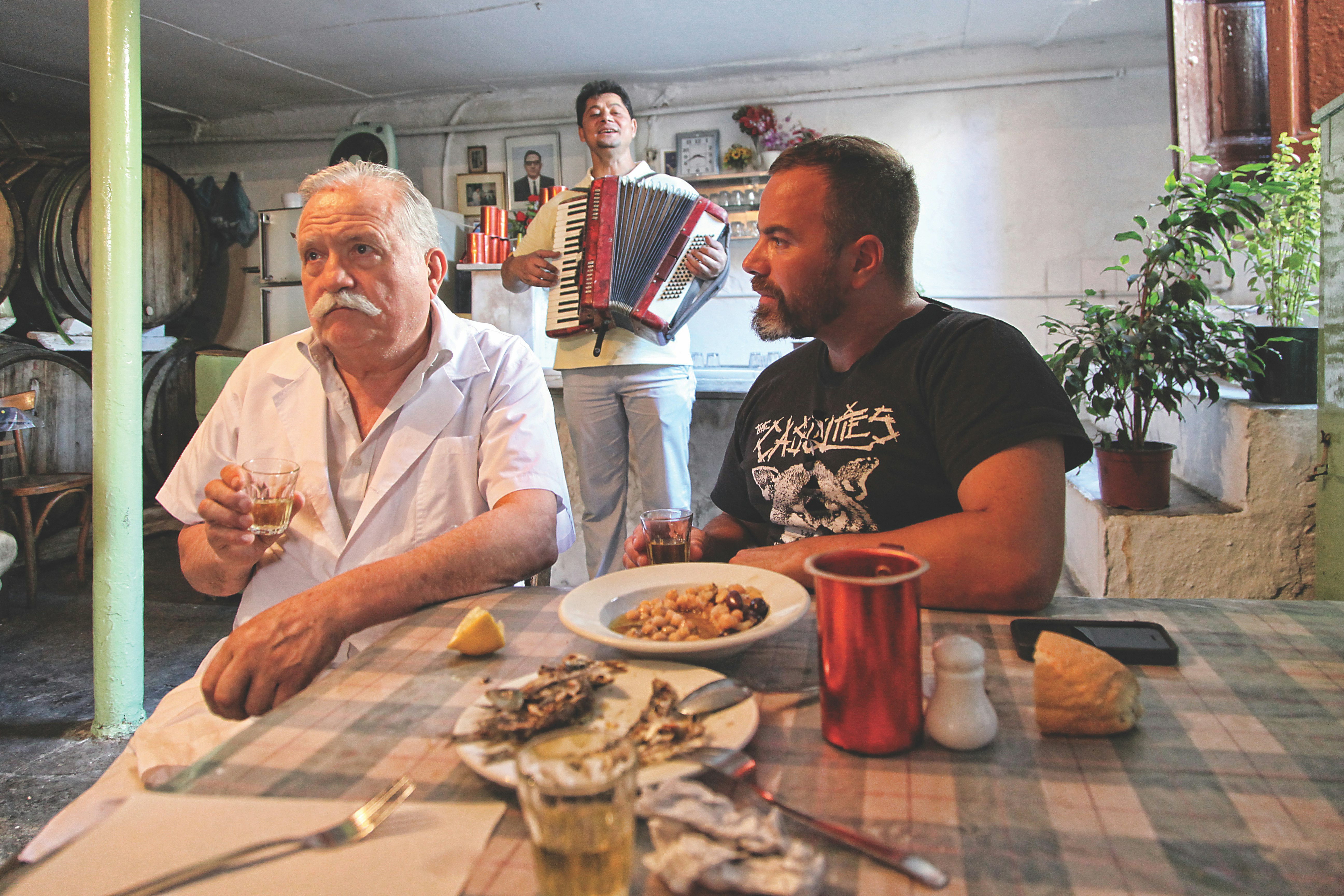 The proprietor of the Diporto taverna in the Monastiraki and Psirri area sits holding a glass of wine. He has a white chefs coat on and a white mustache. To his left sits Kallidis in a black t-shirt with white writing on it. Behind them is an accordion player in white slacks and shirt. On the table are the remains of several dishes and a hunk of bread