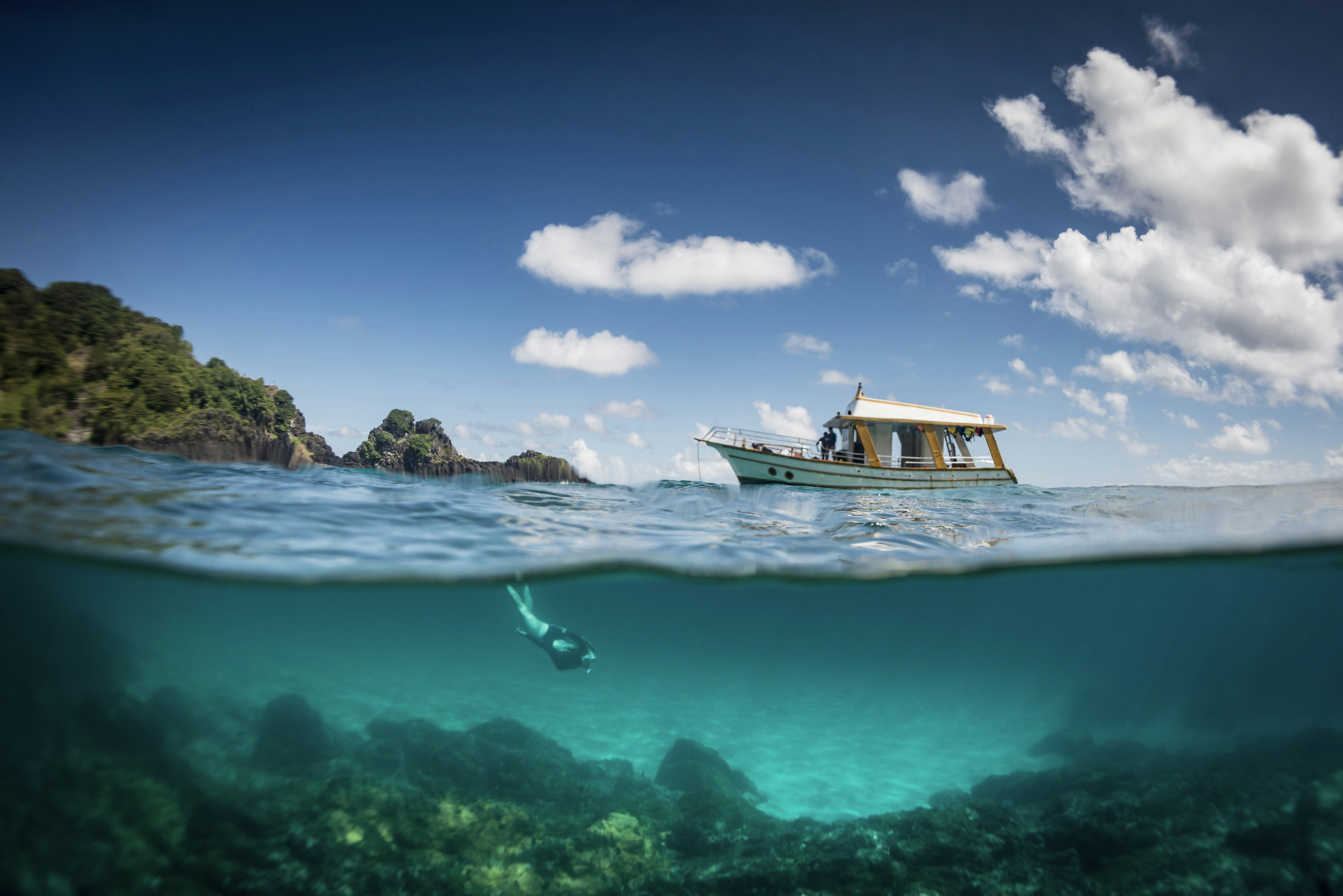 View of a tour boat and a woman free diving at 'Sancho' beach in the archipelago of Fernando de Noronha; the bottom half of the shot shows the diver under water, while the top half shows the boat floating by a rocky shore.