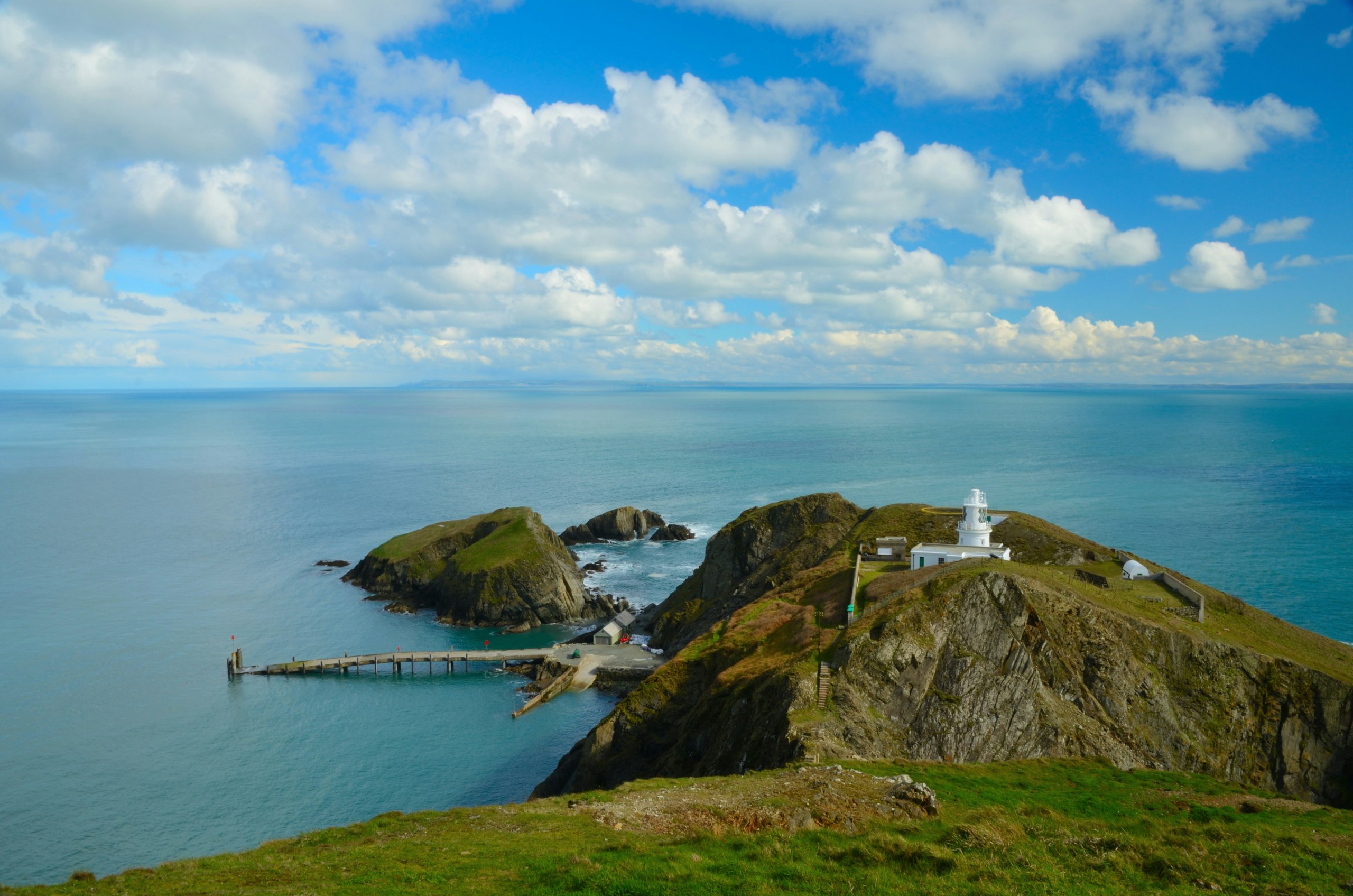 The jetty at Lundy is a narrow wooden walkway jutting out into the sea. There's a white lighthouse tucked among the green clifftops. The mainland can be seen faintly in the background