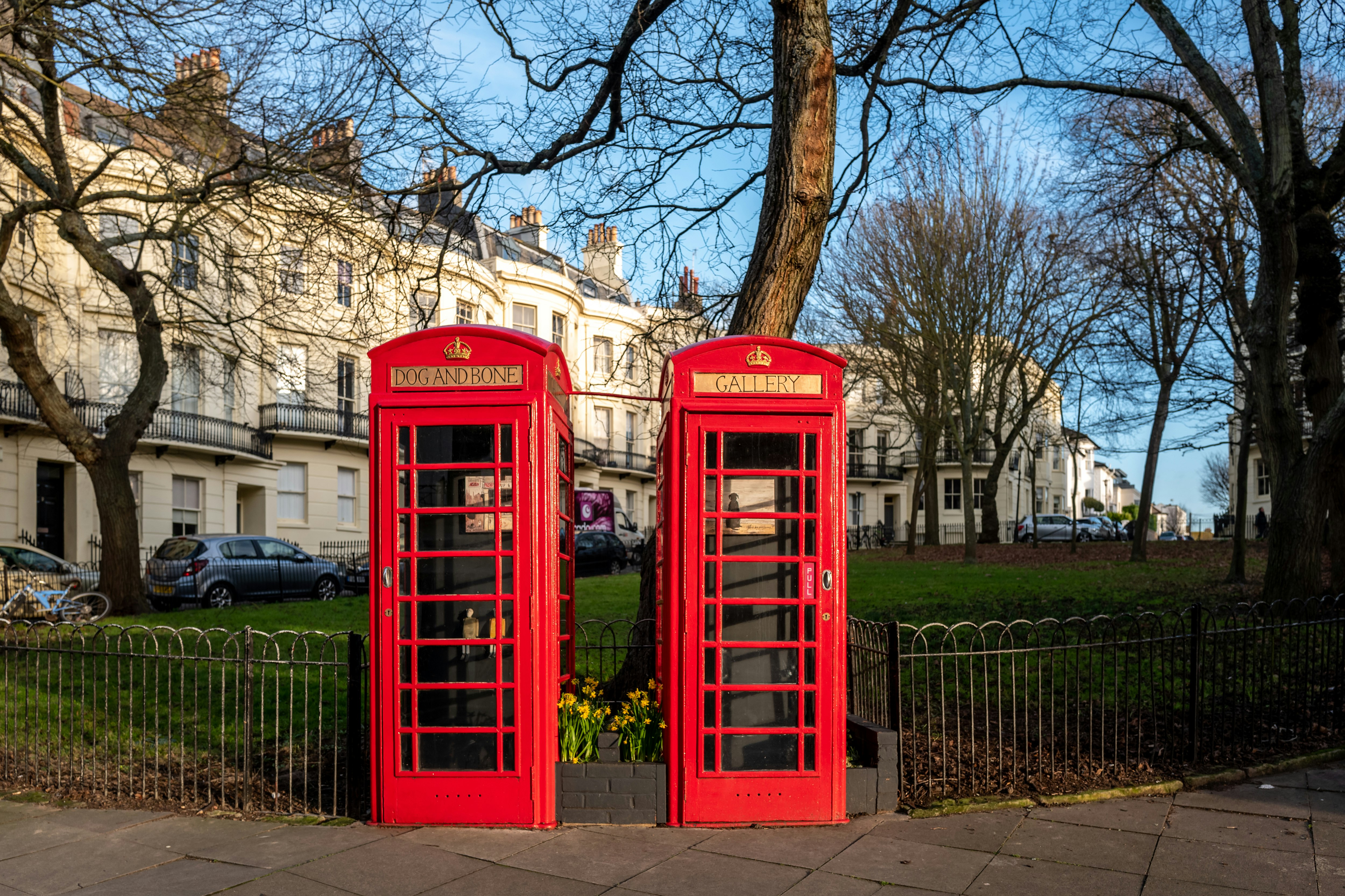 Two traditional red British phone boxes stand side by side on the edge of a green with iron railings around it. In the background is a row of grand white regency-style terraced townhouses.
