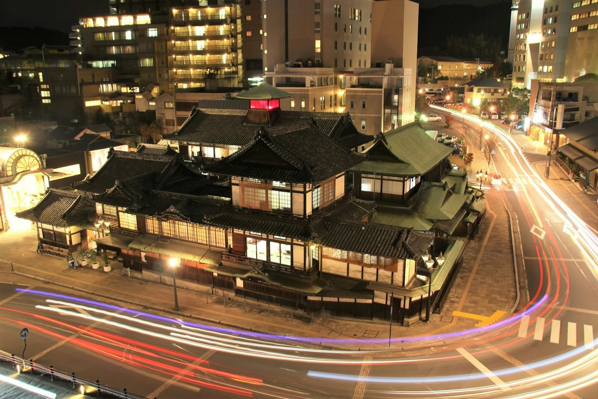A night time shot of the historic Meiji-era sloped, tiled roof of the Dogo Onsen in Ehime Prefecture, with the trails of car headlights and brake lights streaking the road around the structure