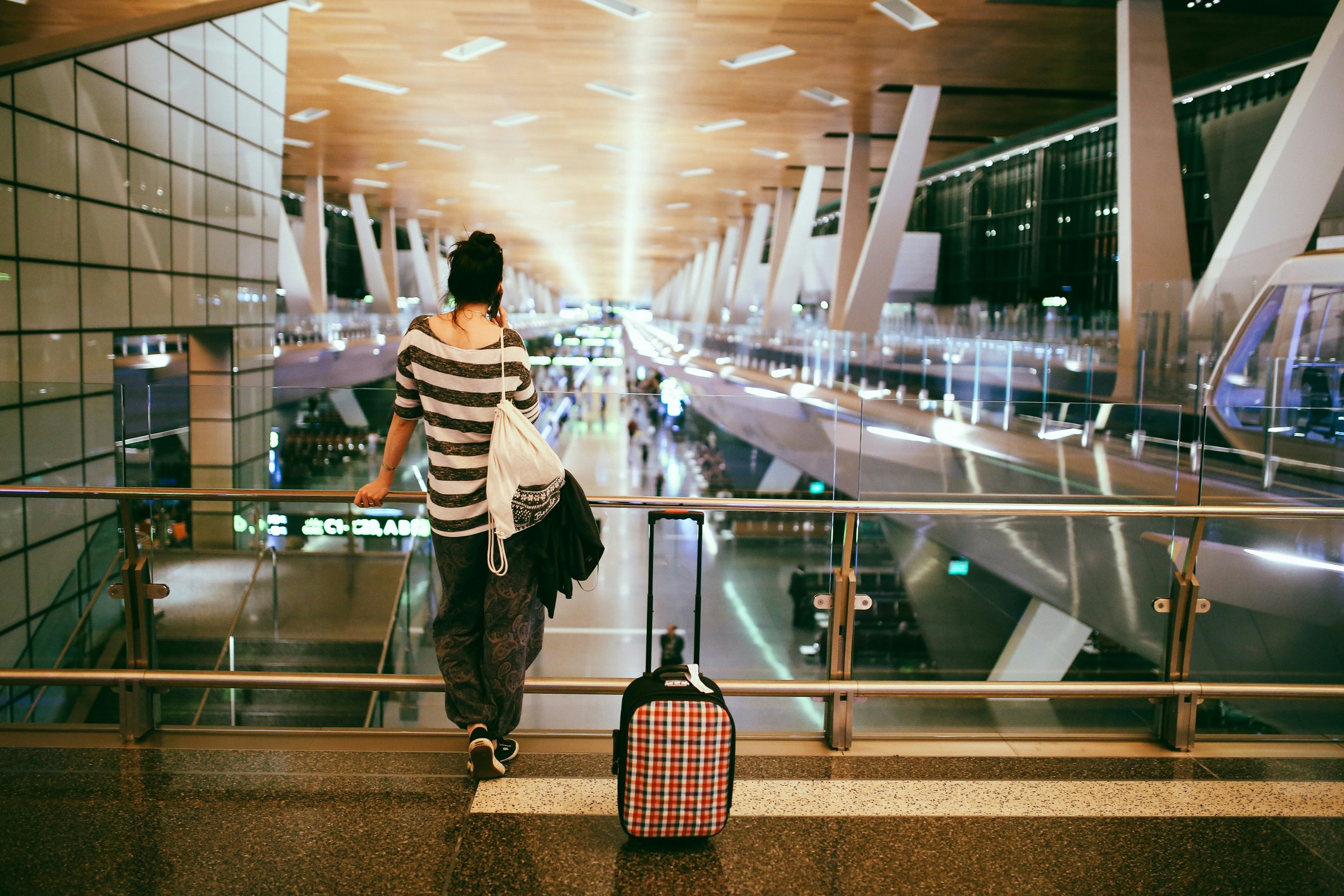 A young woman in baggy grey pants, a grey and white striped shirt, a white athletic-style backpack, and a brightly colored plaid carry-on roller bag stands on the upper level of the Doha airport overlooking the terminal below as she holds a phone to her ear 