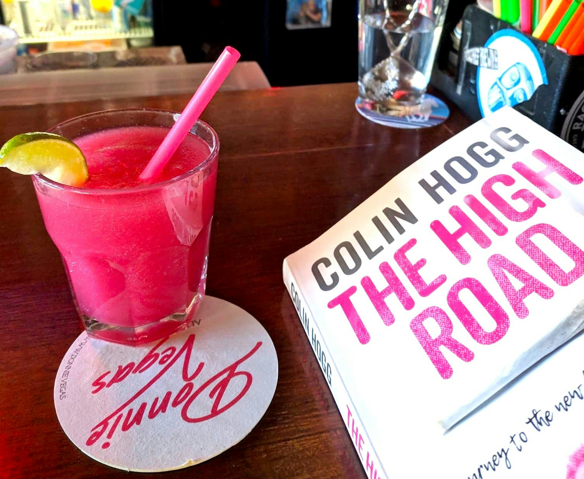 A hot pink cocktail garnished with a lime and with an equally pink plastic straw sits in a highball glass on a coaster reading Donnie Vegas upside down in a fluorescent cursive script. On the right side of the frame is a paperback book by Colin Hogg entitled "The High Road" in a matching pink font 