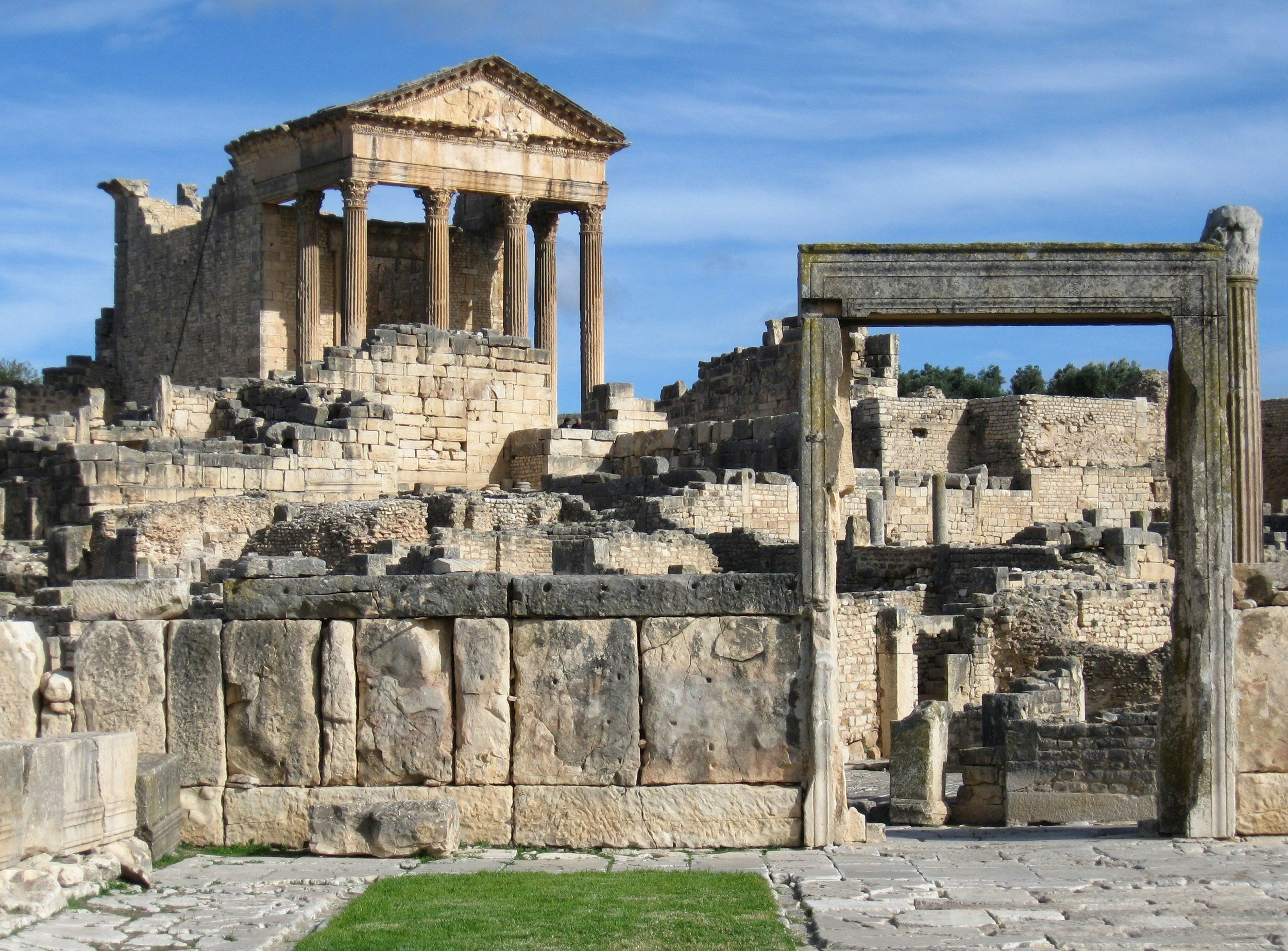 Looking very much like the Forum in Rome, this image is filled with stone ruins; rising into the blue sky at the back of the image is Roman temple with its front facade still held high by six large columns.