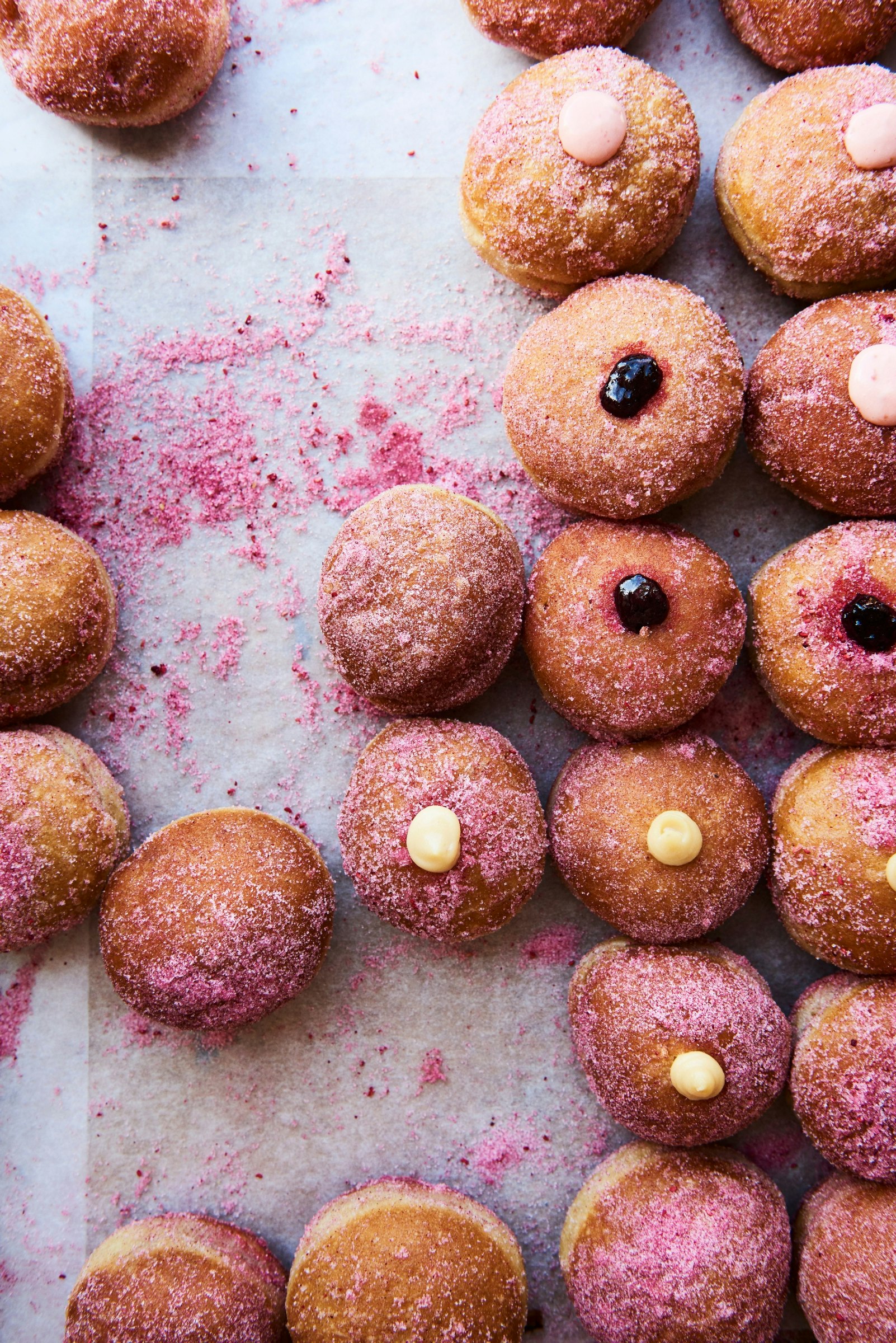 An assortment of sugared doughnuts with blobs of dark-red jam protruding from some and or swirls of yellow custard oozing out of others are lined-up vertically, with a trace of fallen sugar granules showing where other doughnuts would have stood but have now gone 